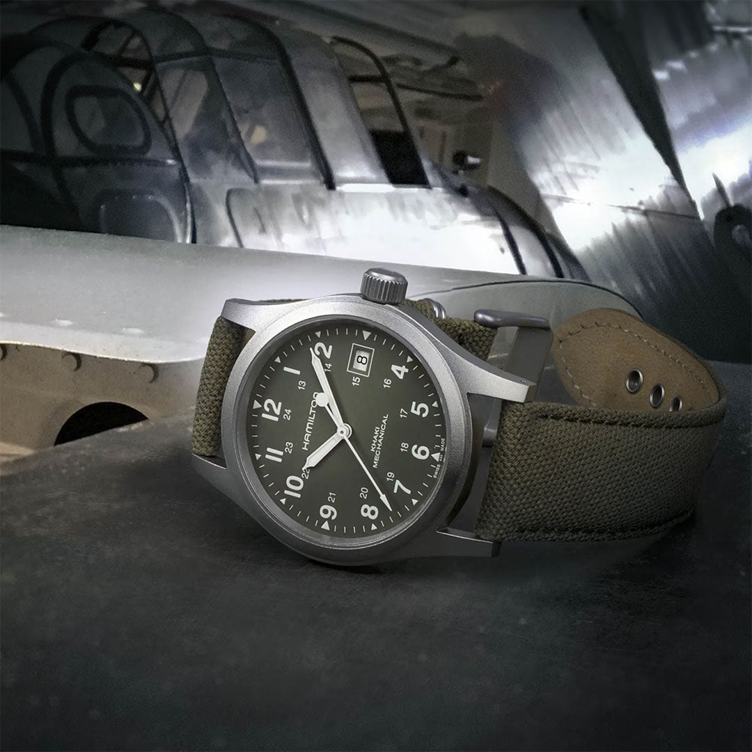 A Hamilton Khaki Field Mechanical 38mm Watch with an olive green strap rests on a surface, while a metallic vintage aircraft appears blurred in the background. Evoking the classic style of the Khaki Field Officer Handwinding, this Hamilton Watch features white numerals and a date display on its dial.