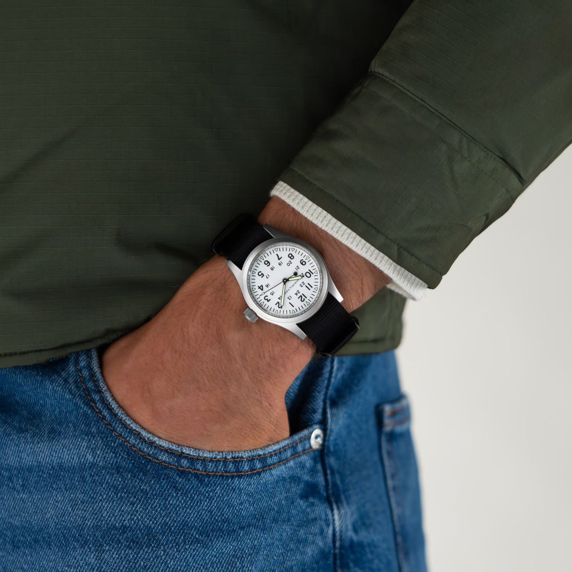 A person in a green jacket and blue jeans, left hand in pocket, wears a Hamilton Khaki Field Mechanical 38mm Watch by Hamilton Watch with a stainless steel case and classic NATO strap.