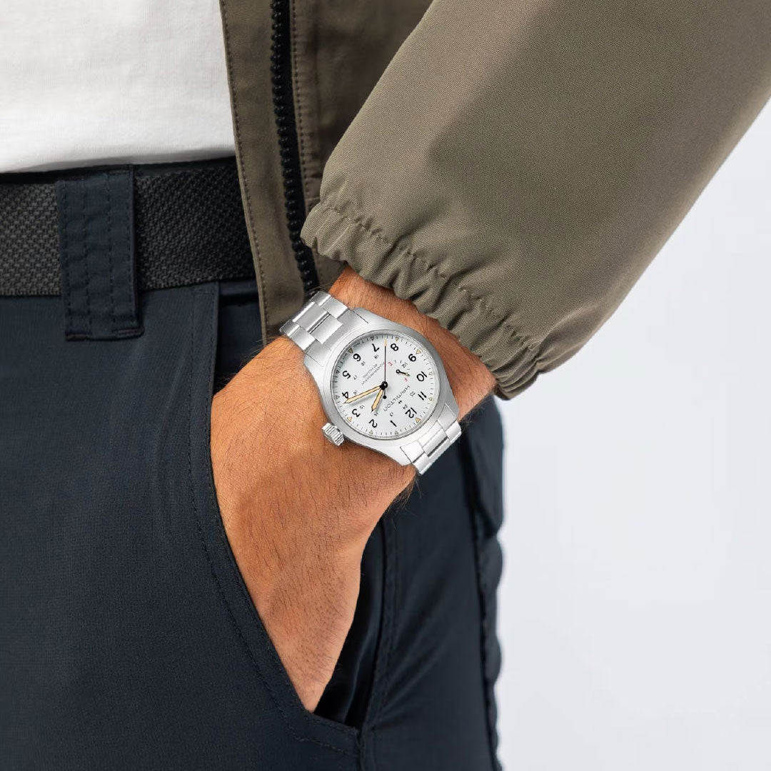 Wearing an olive-green jacket and dark pants, a person slips their hand into their pocket while sporting the Hamilton Khaki Field Mechanical Power Reserve 40mm Watch with a white dial and black numerals.