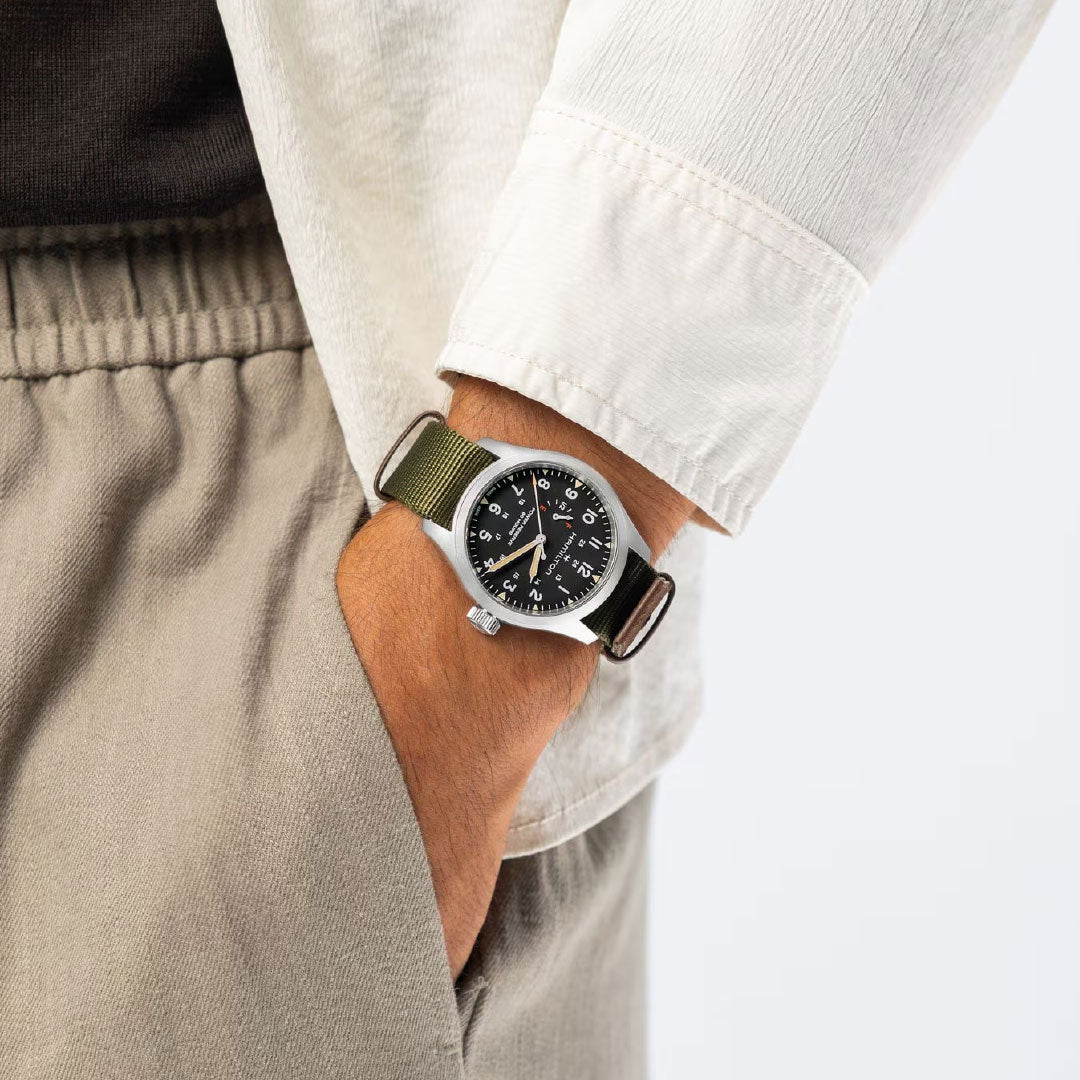 Wearing beige pants and a cream shirt, a person shows off the Hamilton Khaki Field Mechanical Power Reserve 40mm Watch with a black dial, green fabric strap, manual-winding movement, and an 80-hour power reserve by Hamilton Watch.