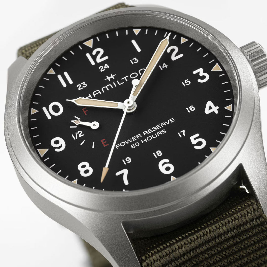 A close-up of the Hamilton Khaki Field Mechanical Power Reserve 40mm watch by Hamilton Watch, featuring a black dial with white numerals, beige hands, an 80-hour power reserve indicator, silver case, and olive green fabric strap.
