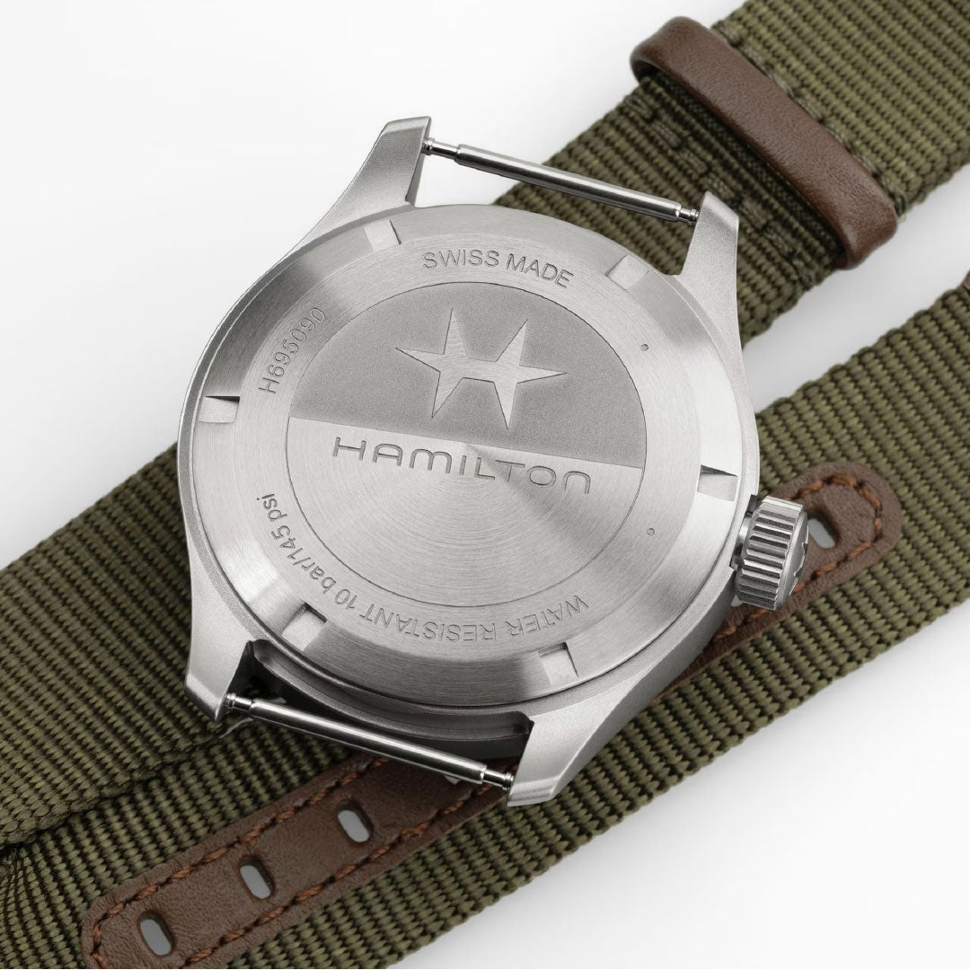 A close-up of the Hamilton Khaki Field Mechanical Power Reserve 40mm Watch by Hamilton Watch, showing its brushed metal case, engraved logo, "SWISS MADE," "WATER RESISTANT TO 10 BAR/145 PSI" on a green fabric strap with brown leather trim.