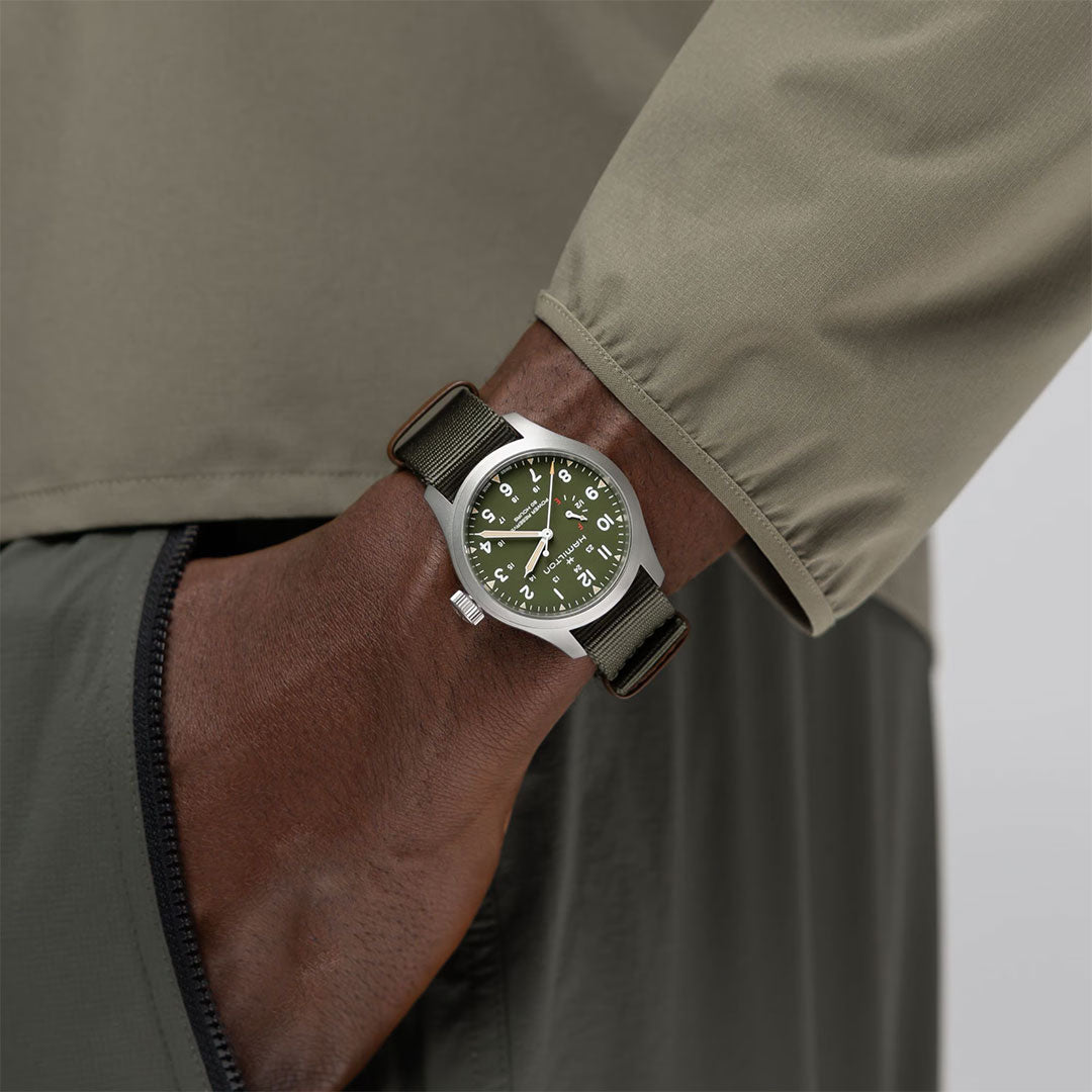 A person in a green jacket has their hand in their pocket, wearing the Hamilton Khaki Field Power Reserve Mechanical 40mm Watch by Hamilton Watch, featuring an olive green dial, fabric strap, and silver case.