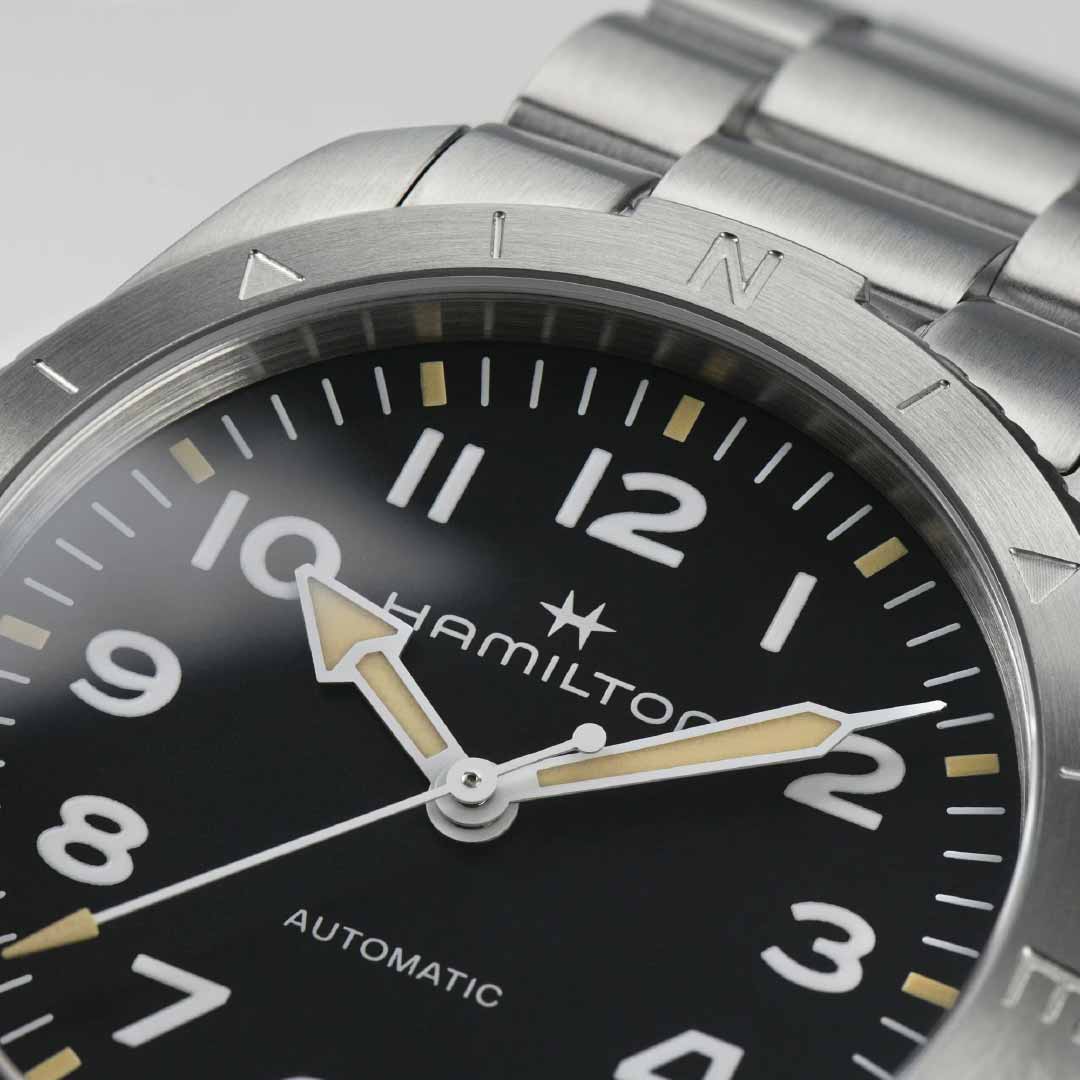 Close-up of a Hamilton Khaki Field Expedition Automatic 41mm Watch by Hamilton Watch, featuring a stainless steel band. The watch's black dial includes white Arabic numerals and luminous hands, capturing the adventurous spirit of the design. The word "Automatic" is printed above the 6 o'clock position.