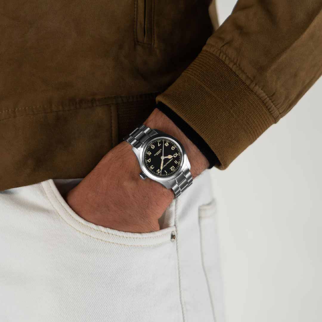 A person sporting a vintage look is clad in a brown suede jacket with white pants, casually resting their hand in their pocket. They wear a Hamilton Khaki Field Murph Automatic 38mm watch by Hamilton Watch, featuring a black dial with striking yellow numerals.