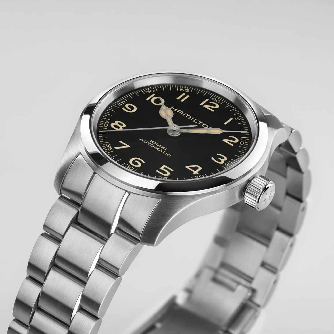 The Hamilton Khaki Field Murph Automatic 38mm Watch, crafted by Hamilton Watch, features a stainless steel case with a black dial adorned with Arabic numerals, ideal for fans of the brand. The watch includes a metal link bracelet and a visible crown, while silver hands with a luminous coating enhance its sophisticated design against a plain light gray background.