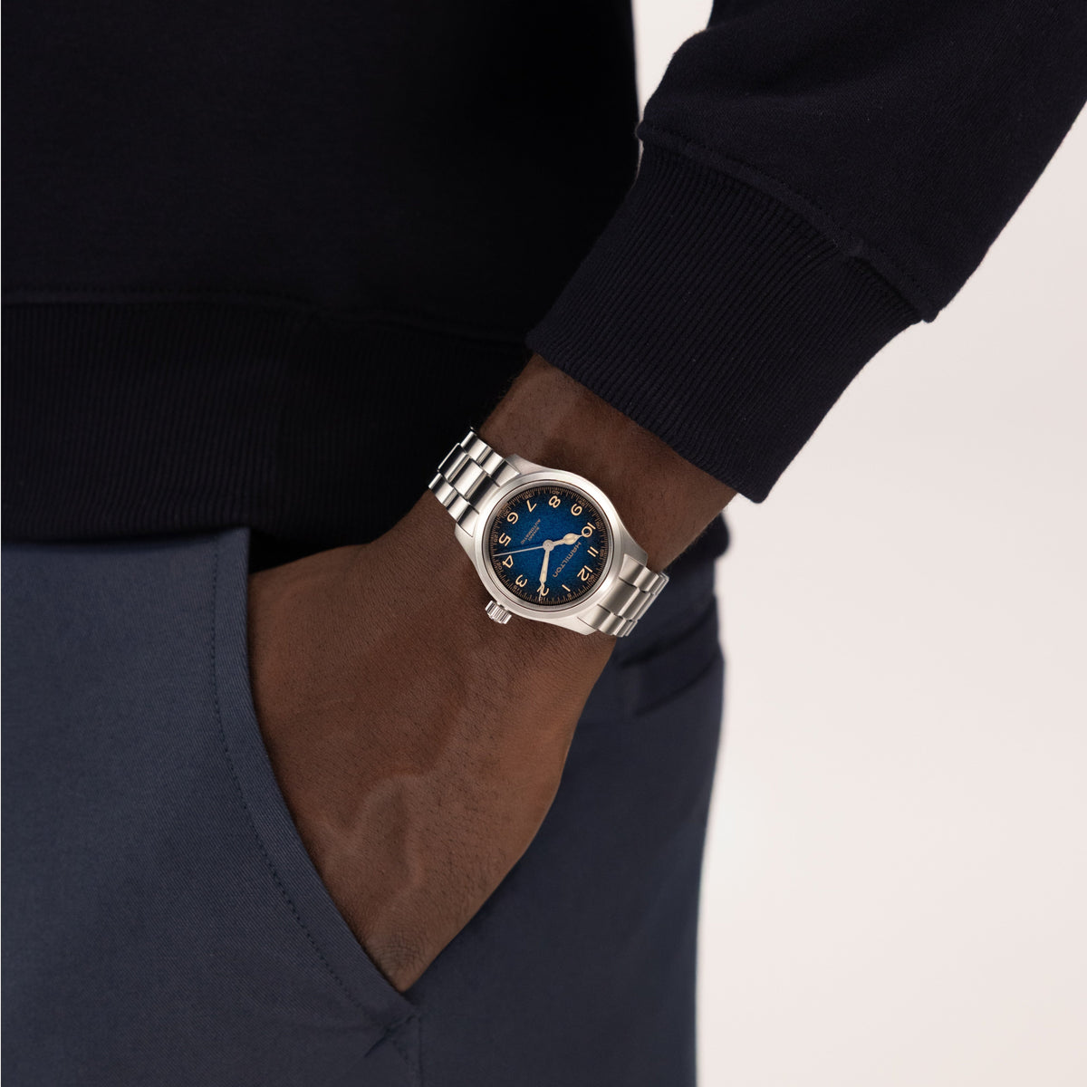 A person wearing a Hamilton Khaki Field Murph Auto 38mm Watch by Hamilton Watch, featuring a striking blue-to-black dial and silver case, has their hand tucked into the pocket of dark blue pants. Dressed in a dark long-sleeve top, they pair refined style with the precision craftsmanship of this automatic calibre timepiece.