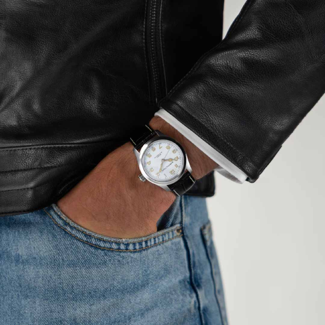 A person wearing a black leather jacket and blue jeans, with their hand in the pocket. They sport a vintage timepiece, the Hamilton Khaki Field Murph Automatic 38mm Watch by Hamilton Watch, featuring a silver casing with a white face and black leather strap.