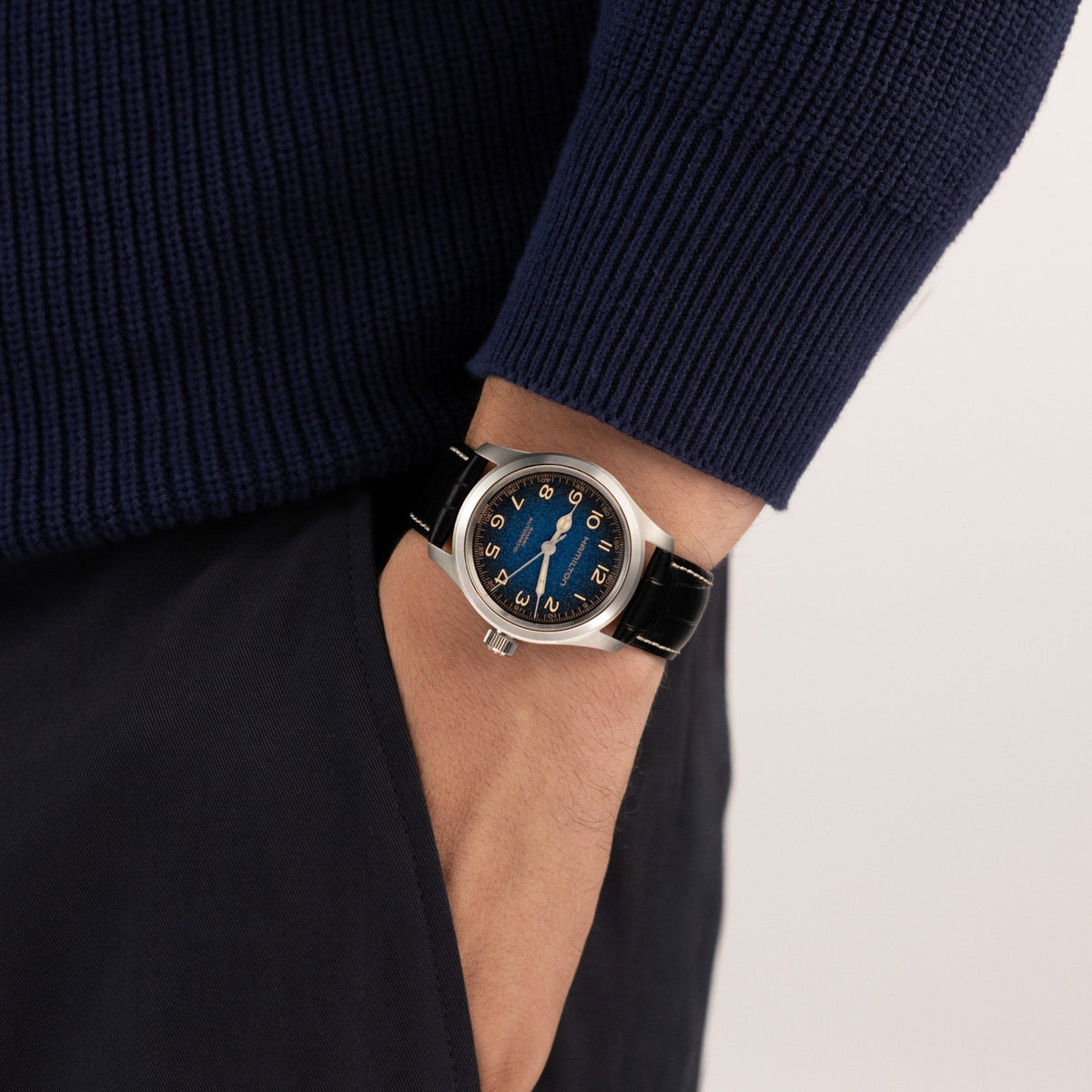 A person dressed in a navy sweater and dark pants has their hand in their pocket, showcasing the Hamilton Khaki Field Murph Auto 38mm Watch by Hamilton Watch, which features a striking blue-to-black dial with gold numerals and a black leather strap.