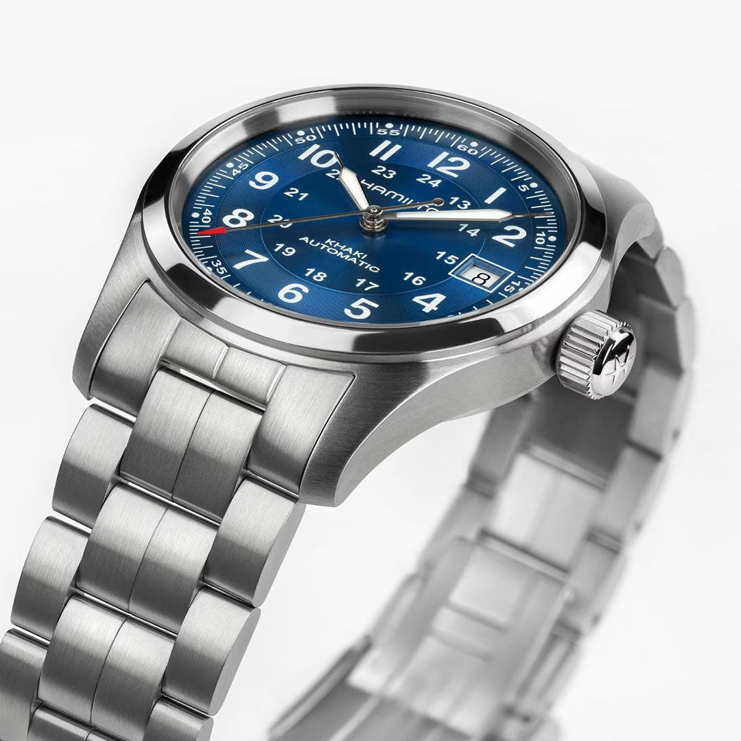 A close-up of the Hamilton Watch Khaki Field Auto 38mm shows its blue dial with white numerals, date window, and metal link bracelet, reflecting military heritage. The time is set to 10:10.
