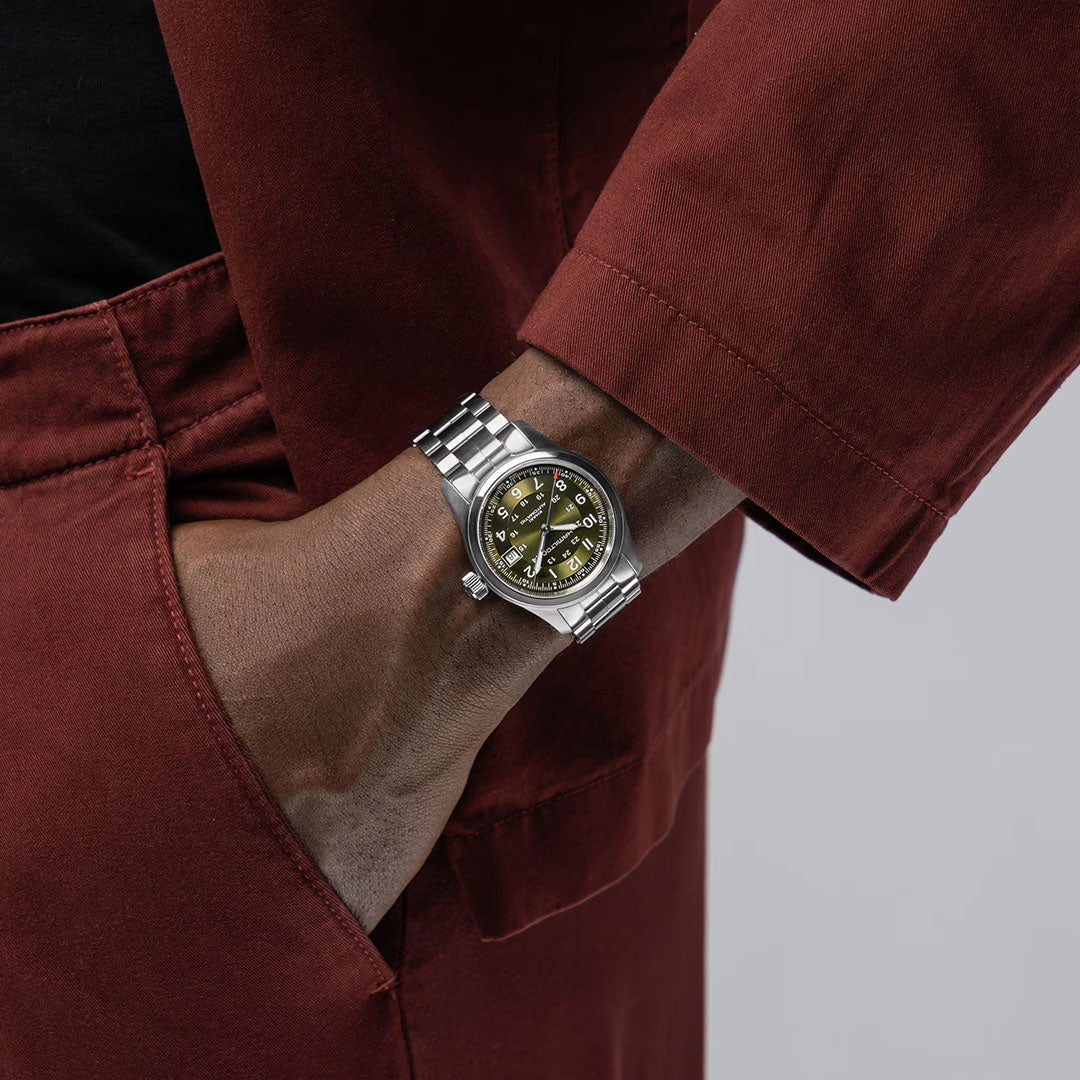 A person in a red jacket with their hand in their pocket, wearing a Hamilton Khaki Field Auto 38mm Watch by Hamilton Watch, featuring a green face, silver case, metal band, and automatic movement.