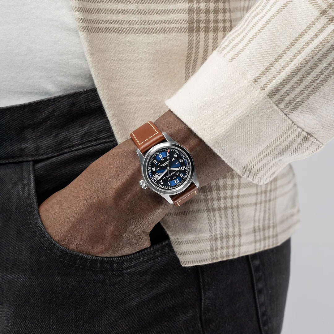 A person in a beige plaid shirt and black jeans shows off a Hamilton Khaki Field Auto 38mm Watch from Hamilton Watch, featuring a blue dial, brown leather strap, and the signature H-10 automatic movement inspired by military heritage.