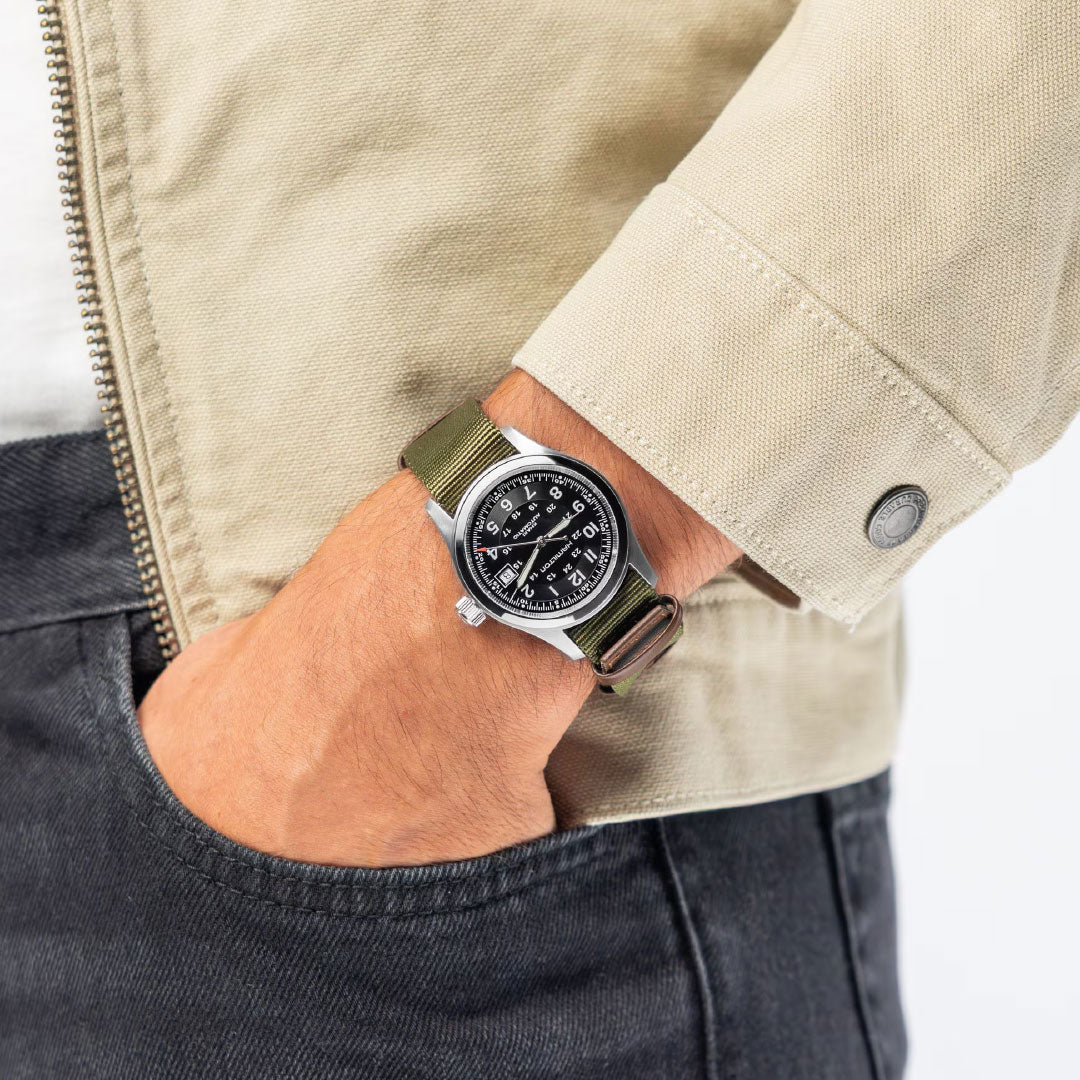 A person in a beige jacket and dark jeans, left hand in their pocket, wears a Swiss-made Hamilton Khaki Field Auto 38mm Watch Call of Duty with a black dial and olive green strap by Hamilton Watch.