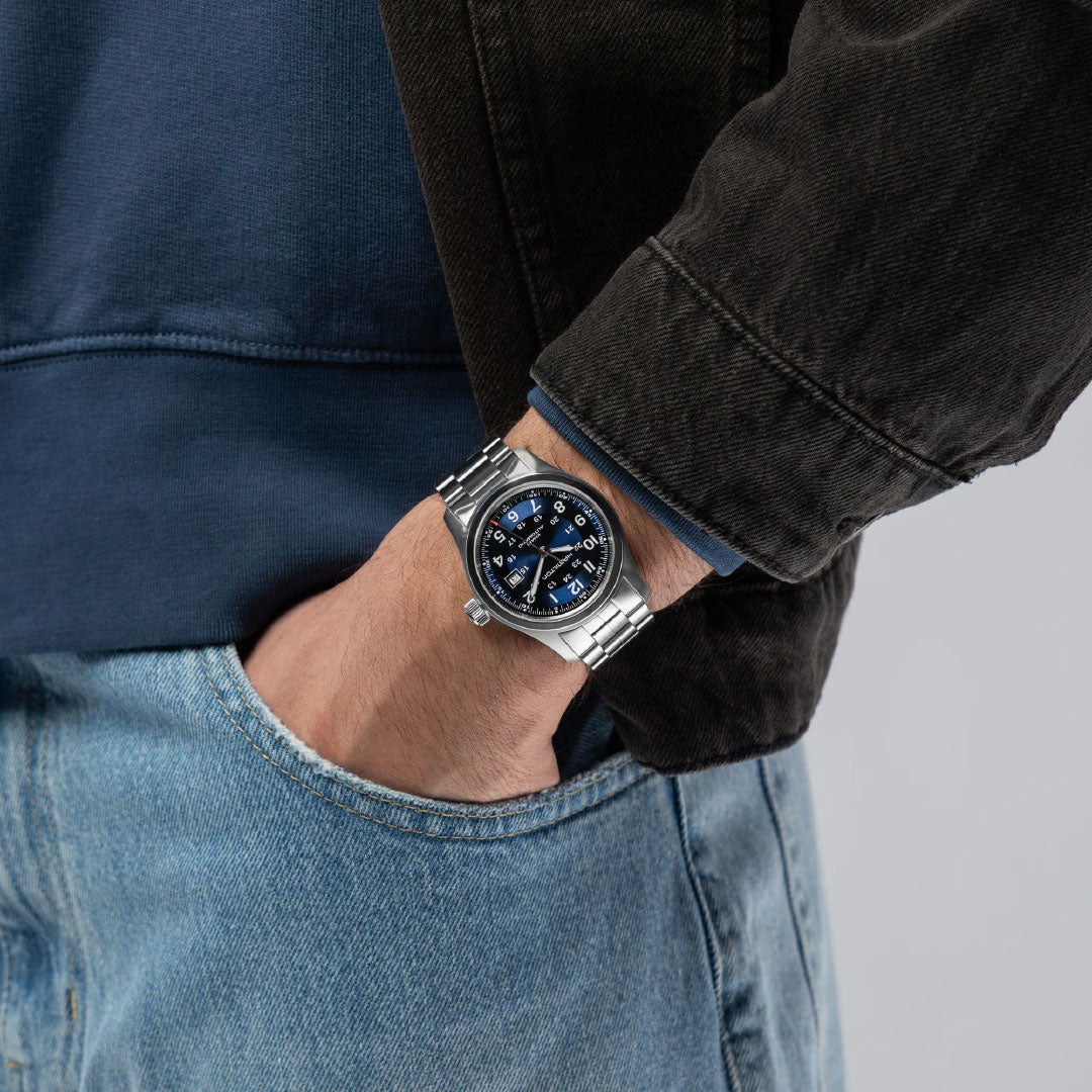 A person in a dark jacket and blue jeans has their left hand in their pocket, displaying the Hamilton Khaki Field Auto 42mm Watch—an automatic timepiece by Hamilton Watch that echoes classic military heritage with its striking blue face and silver accents.