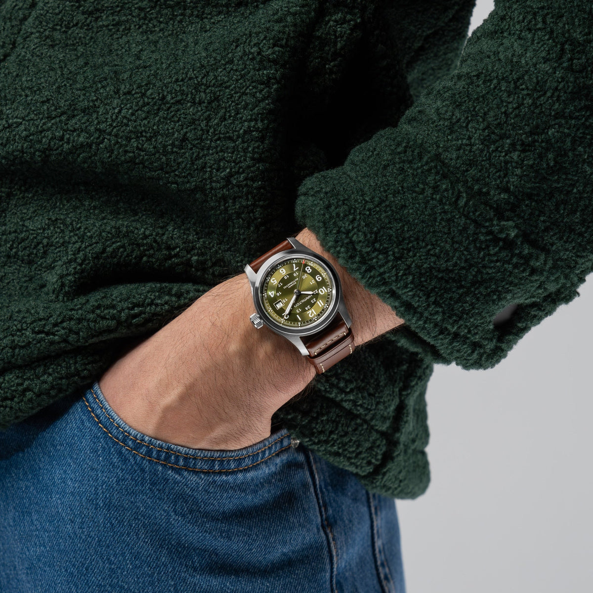A person dressed in a green fleece jacket and blue jeans has their left hand in their pocket, displaying a Hamilton Watch Khaki Field Auto 42mm with a green dial and brown leather strap.