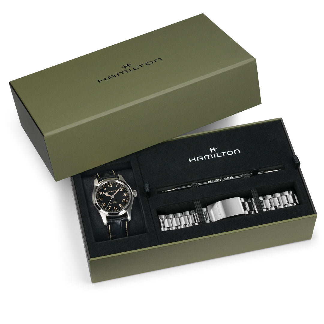 A green Hamilton Watch box with the lid partially open reveals the Hamilton Khaki Field Murph Auto 42mm Watch Bracelet Bundle, including a black analog watch, extra bracelet links, and an adjustment tool inside.
