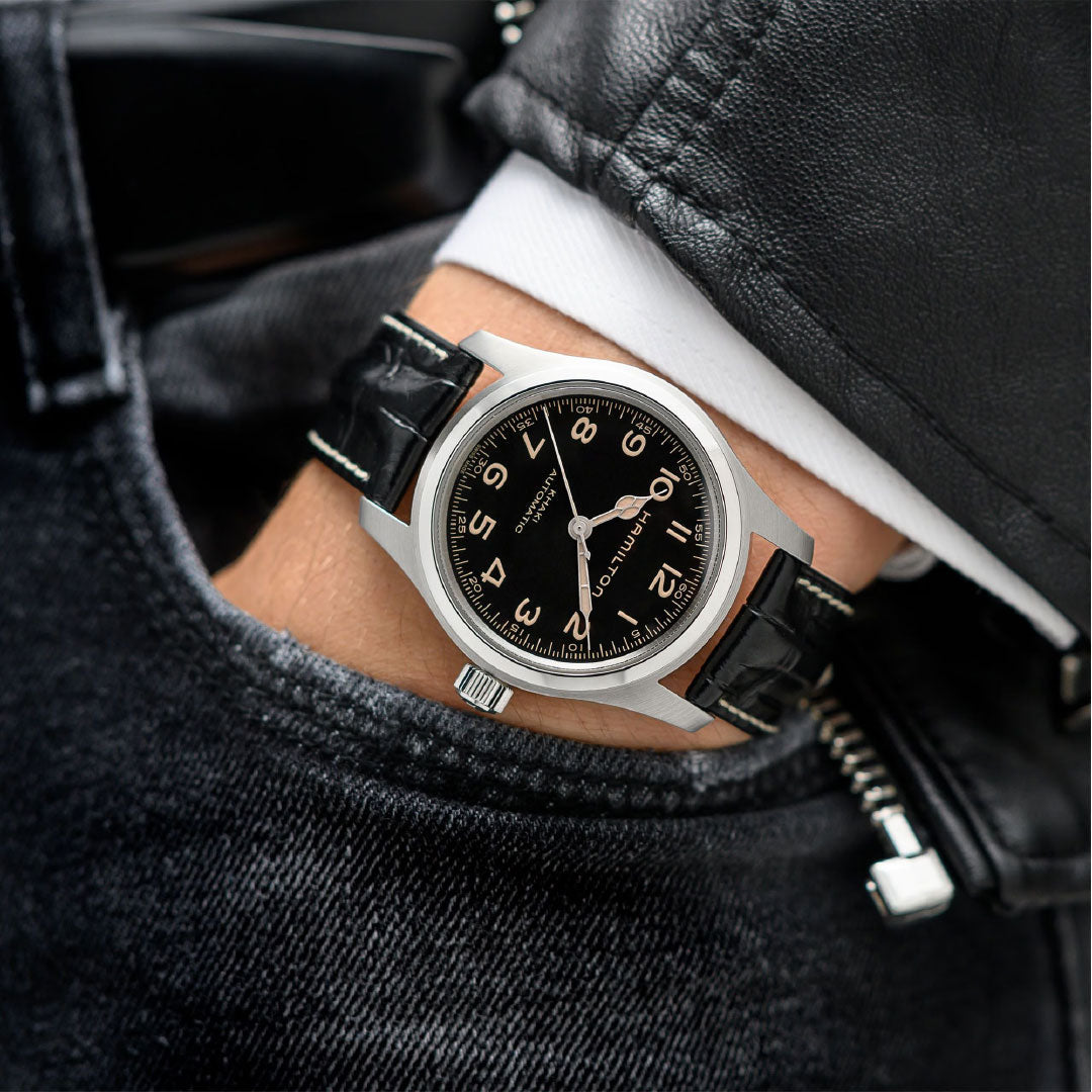 A person wearing a black leather jacket has their hand in a pocket, showcasing the Hamilton Khaki Field Murph Auto 42mm Watch Bracelet Bundle by Hamilton Watch, featuring a classic black dial with white Arabic numerals.