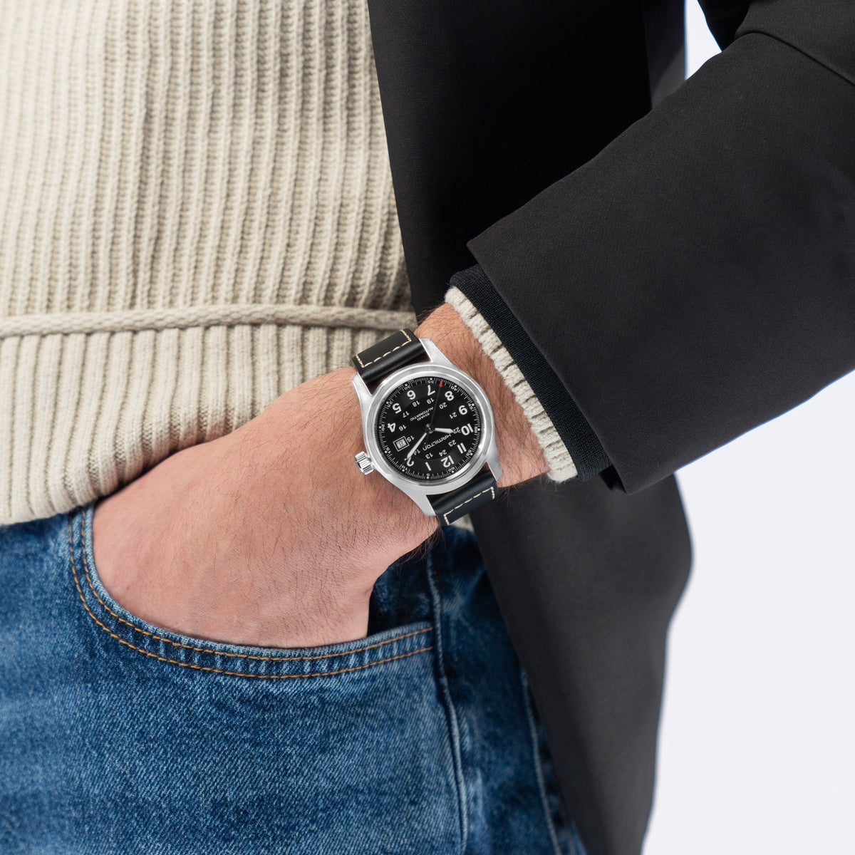 A person wearing a cream ribbed sweater, dark blazer, and blue jeans has their left hand in their pocket and is sporting a Hamilton Khaki Field Auto 42mm Watch by Hamilton Watch on their wrist.