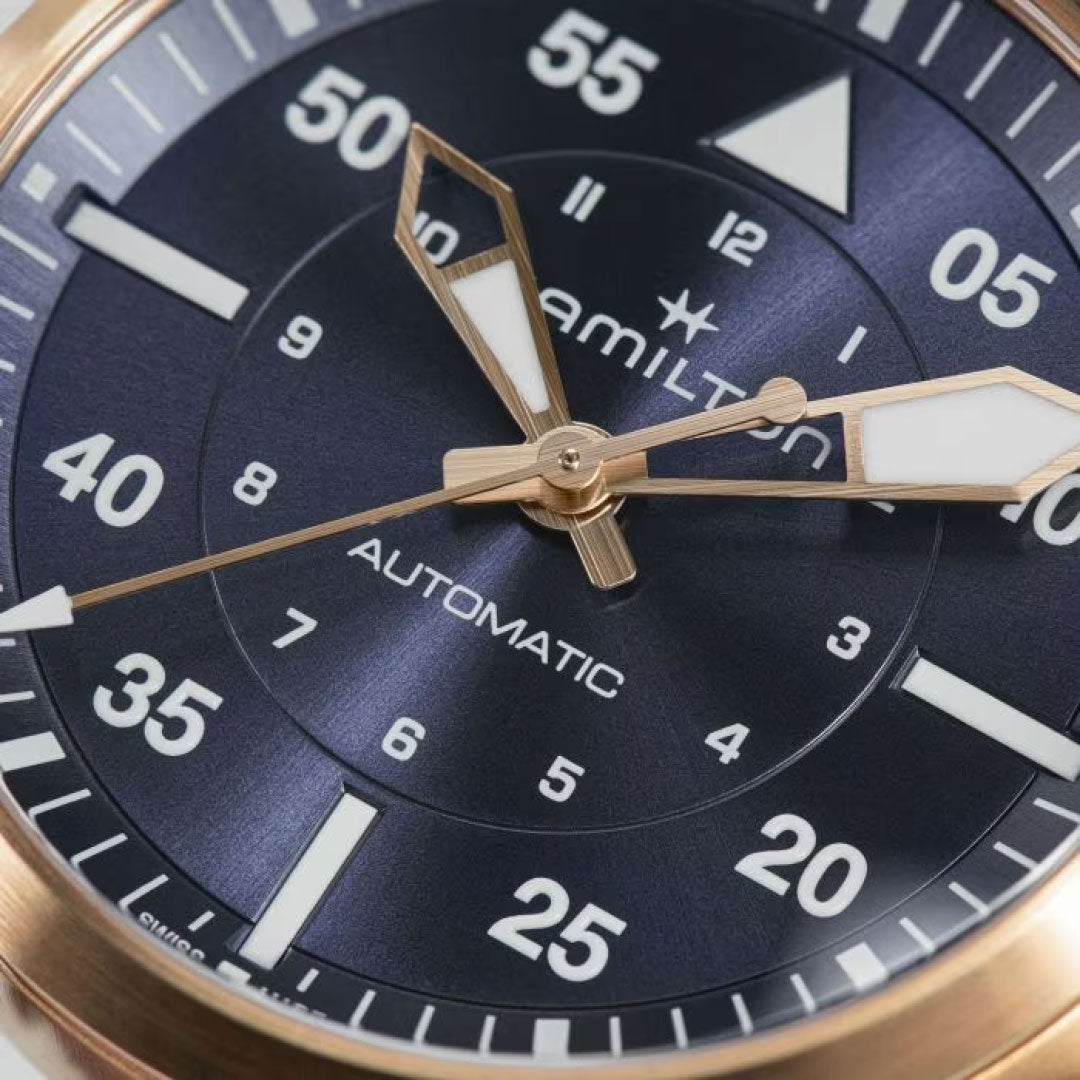 Close-up image of the Hamilton Khaki Aviation Pilot Auto 36mm Watch showcasing a dark blue analog watch face. It features gold-tone hands and white hour markers coated with Super-LumiNova. The watch prominently displays minutes in increments of five, and includes text such as "Hamilton" and "Automatic.