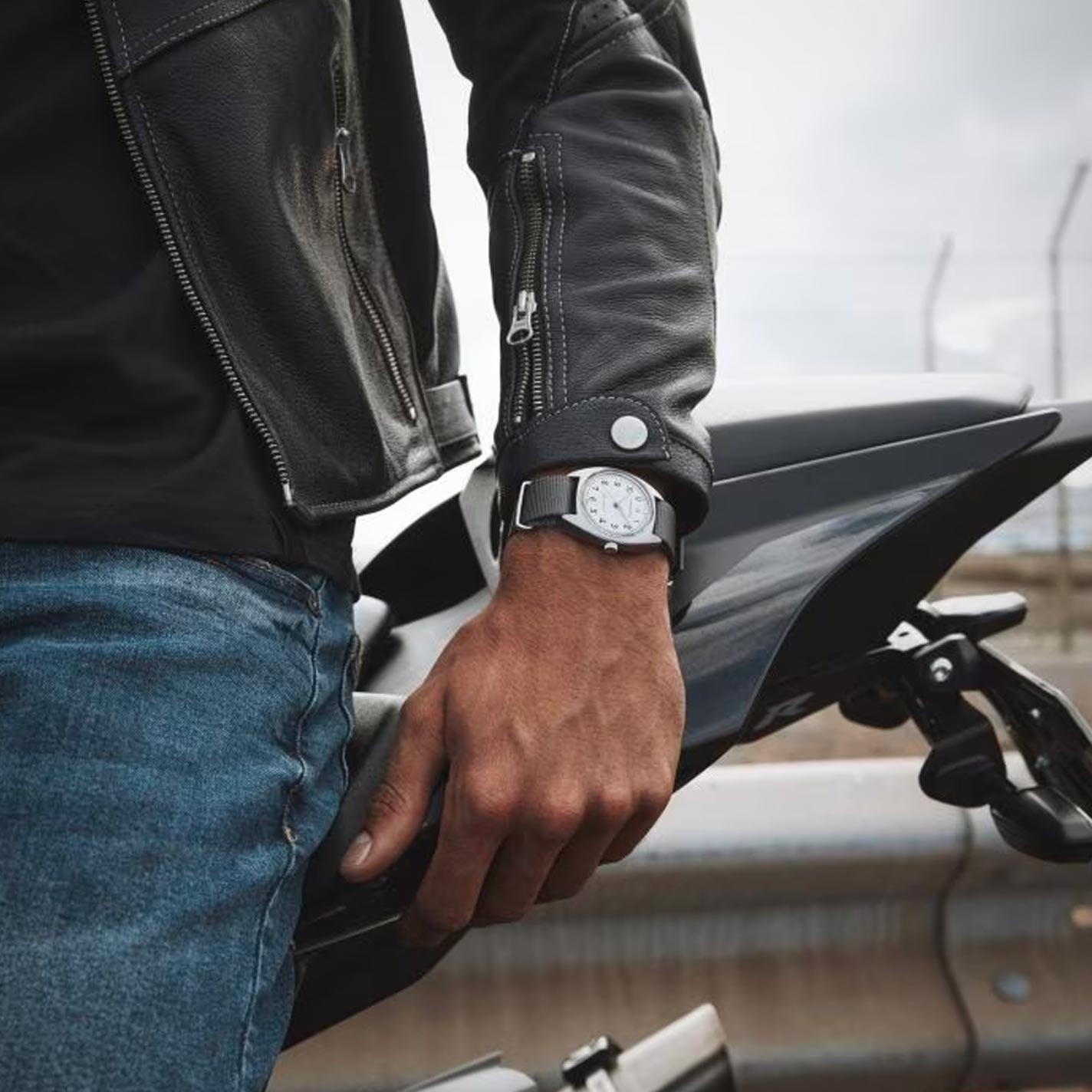 A person in a black leather jacket and jeans leans on a motorcycle, subtly showcasing their Hamilton Khaki Aviation Pilot Pioneer Mechanical 36mm x 33mm Watch. The background features a metal guardrail and an overcast sky, evoking the adventurous spirit associated with Hamilton Watch's legacy.