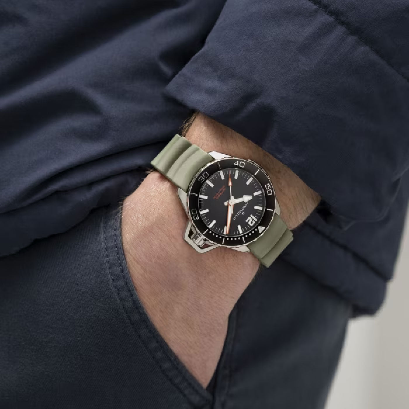 The individual is wearing a dark jacket with one hand in a pocket, while sporting the Hamilton Khaki Navy Frogman Auto 41mm Watch by Hamilton Watch. The wristwatch features a silver case, black bezel, and olive green strap, complemented by a black dial displaying the date function and adorned with a red second hand.