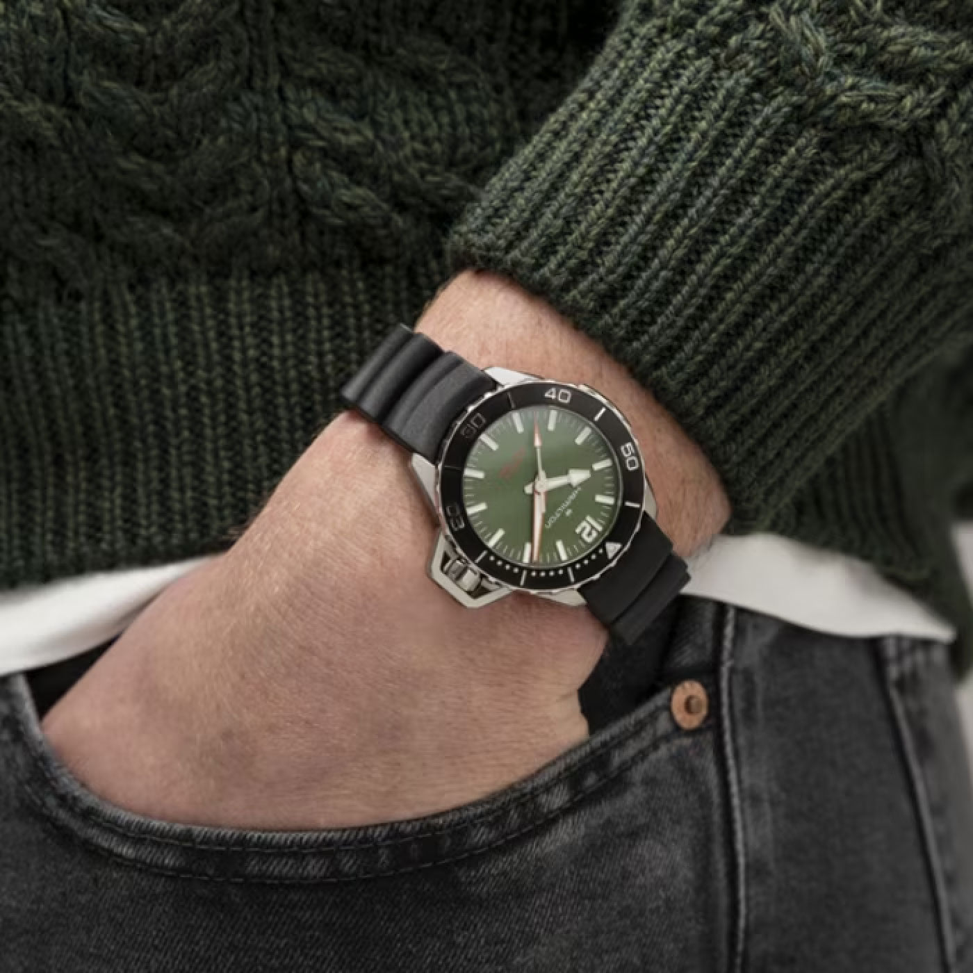 A person in a green sweater and jeans is showcasing the Hamilton Khaki Navy Frogman Auto 41mm Watch on their wrist. This timepiece from Hamilton Watch features a green dial, a black bezel with white markings, and is paired with a sleek black strap.