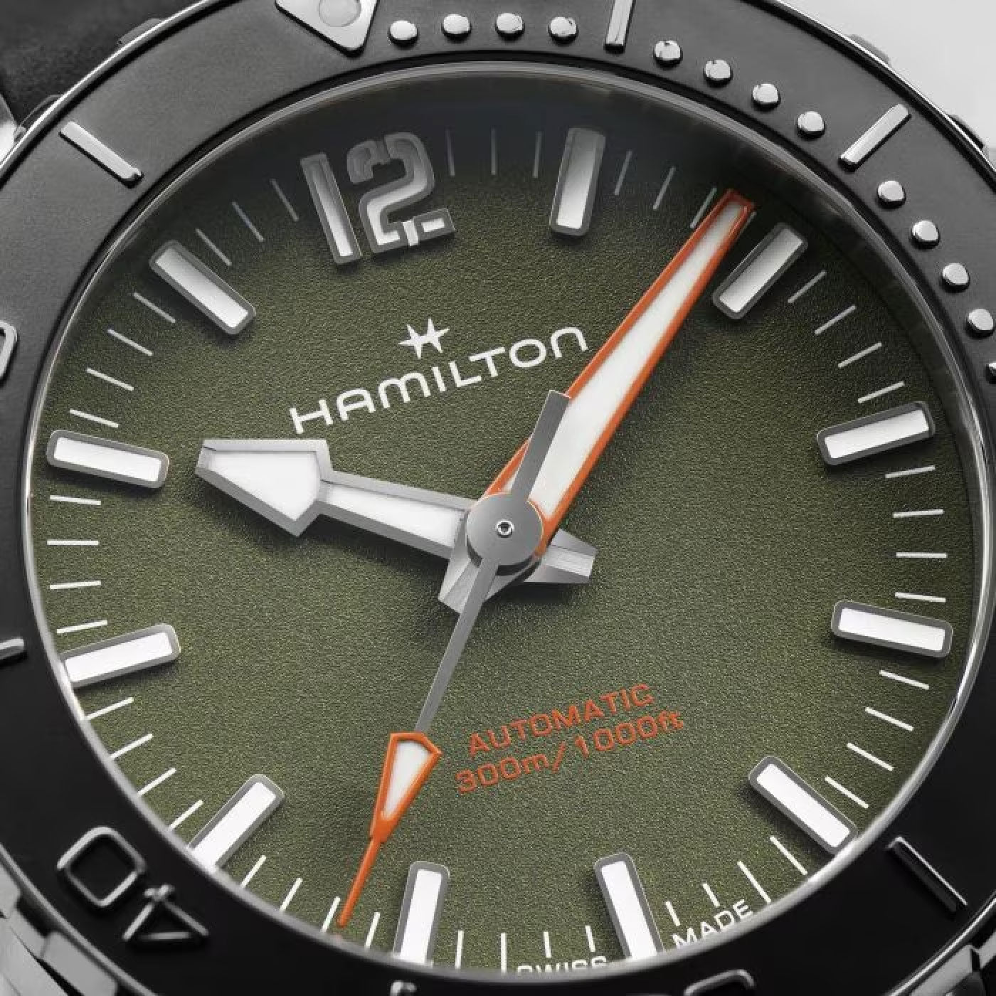 Close-up of the Hamilton Khaki Navy Frogman Auto 41mm watch face showcasing a vibrant green dial, sleek silver hour markers, and white hands accented with orange. The brand "Hamilton Watch" is prominently displayed along with "AUTOMATIC 300m/1000ft" in bold orange text. The time shows approximately 12:02.