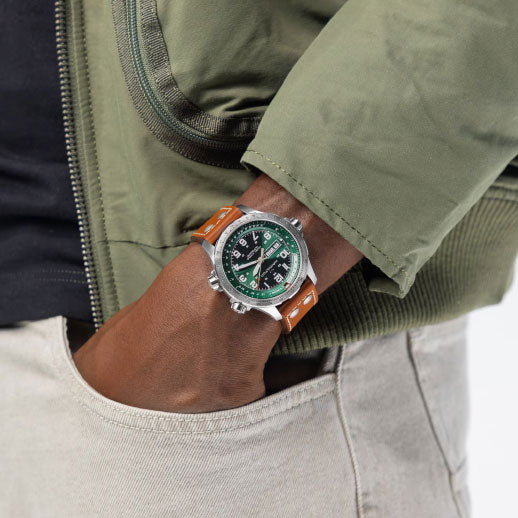A person in a green bomber jacket and light pants has their hand in their pocket, revealing the Hamilton Khaki Aviation X-Wind Day Date Auto 45mm Watch by Hamilton Watch, featuring a green dial and brown leather strap, on their wrist.