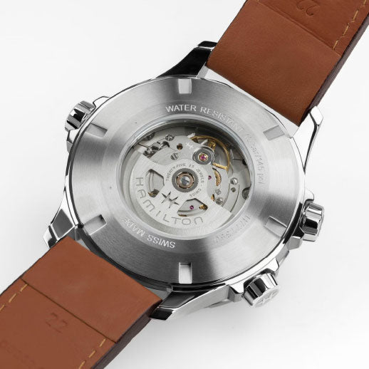 A close-up of the Hamilton Khaki Aviation X-Wind Day Date Auto 45mm Watch reveals its transparent back showcasing the H-30 movement with an 80-hour power reserve, brown leather strap, Swiss made markings, and water resistance.