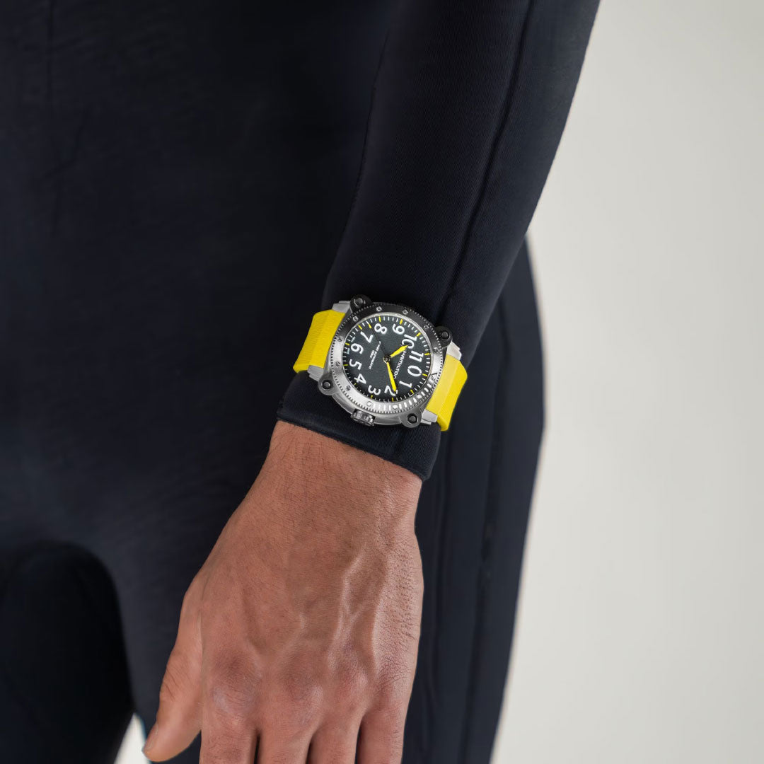 A person in a black wetsuit displays their left hand, wearing the Hamilton Khaki Navy BeLow Zero Auto Titanium 46mm Watch by Hamilton Watch, which features a yellow strap and black-and-silver case, standing out against a plain light background.