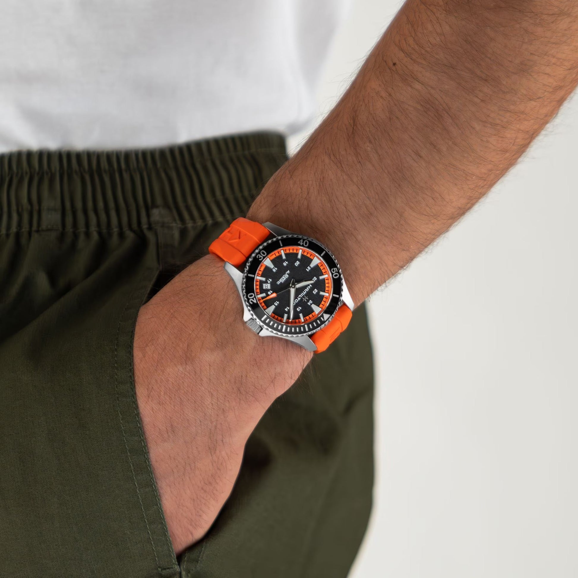 Wearing olive green pants and a white shirt, this person sports the Hamilton Khaki Navy Scuba Auto 40mm Watch by Hamilton Watch, featuring a tangerine orange rubber strap and black dial with white and orange markers.