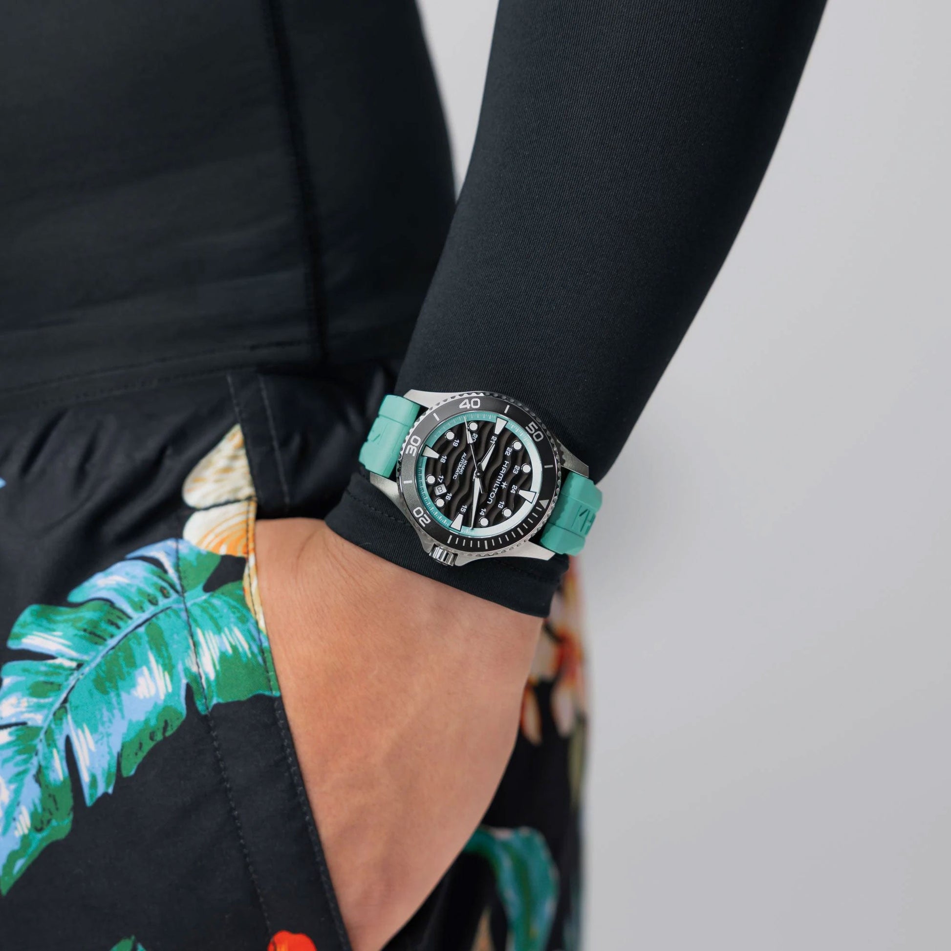 A person in a long-sleeve black shirt and floral shorts, hand in pocket, showcases the Hamilton Khaki Navy Scuba Auto 40mm Watch by Hamilton Watch with a black dial and light turquoise strap.