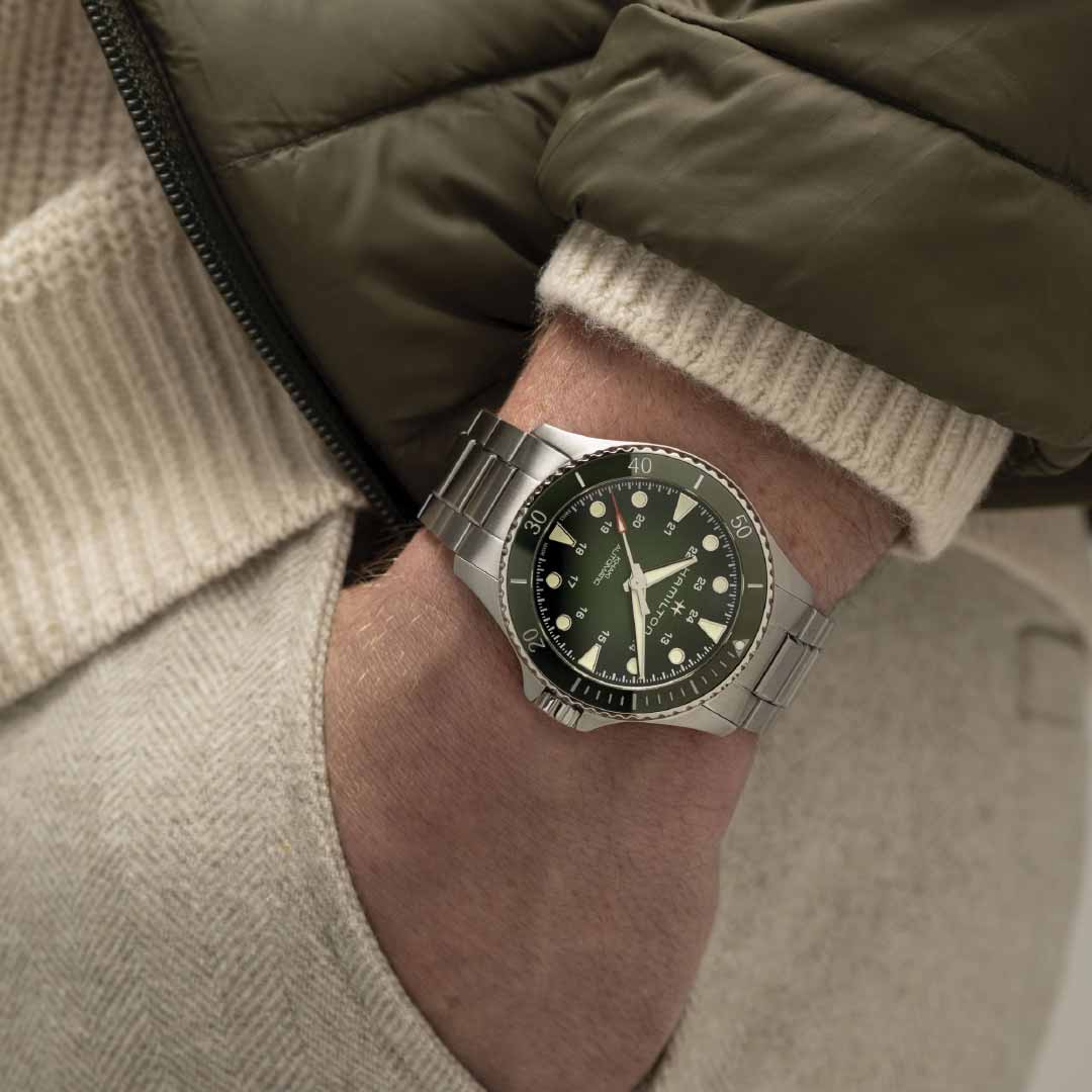 A person is donning the Hamilton Khaki Navy Scuba Automatic 43mm Watch, a sleek silver wristwatch by Hamilton Watch. The watch features a black dial with white markings and is powered by the H-10 automatic movement. Its design includes a green ceramic bezel that subtly emerges from beneath a beige sweater sleeve and an olive-green jacket, as their hand is tucked into the pocket of beige pants.