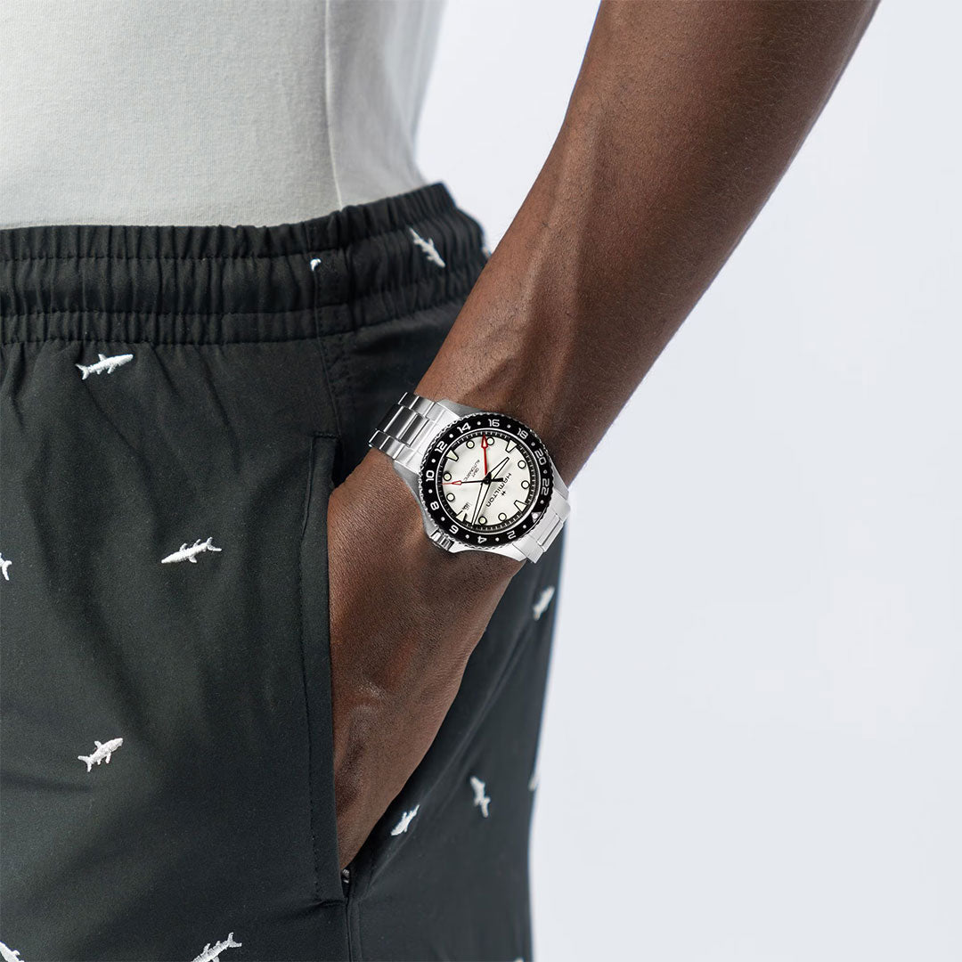 A person in black shorts with white shark patterns, hand in pocket, showcases a Hamilton Khaki Navy Scuba Auto GMT 43mm watch with a black and white bezel on their wrist against a light background.