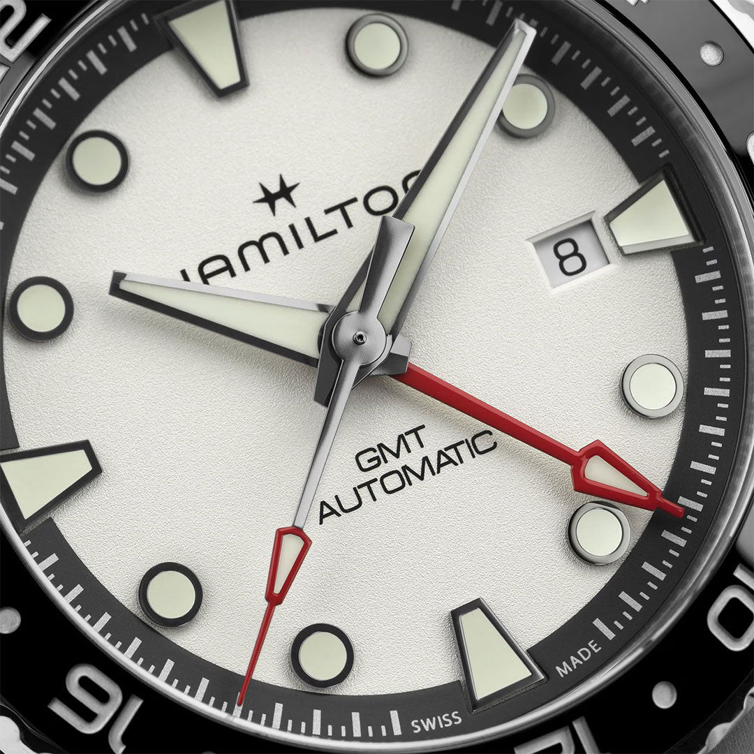 Close-up of a Hamilton Khaki Navy Scuba Auto GMT 43mm Watch face by Hamilton Watch, featuring black and white markers, luminous hands, red GMT hand, date at “8,” and “Hamilton GMT Automatic” on the white dial.