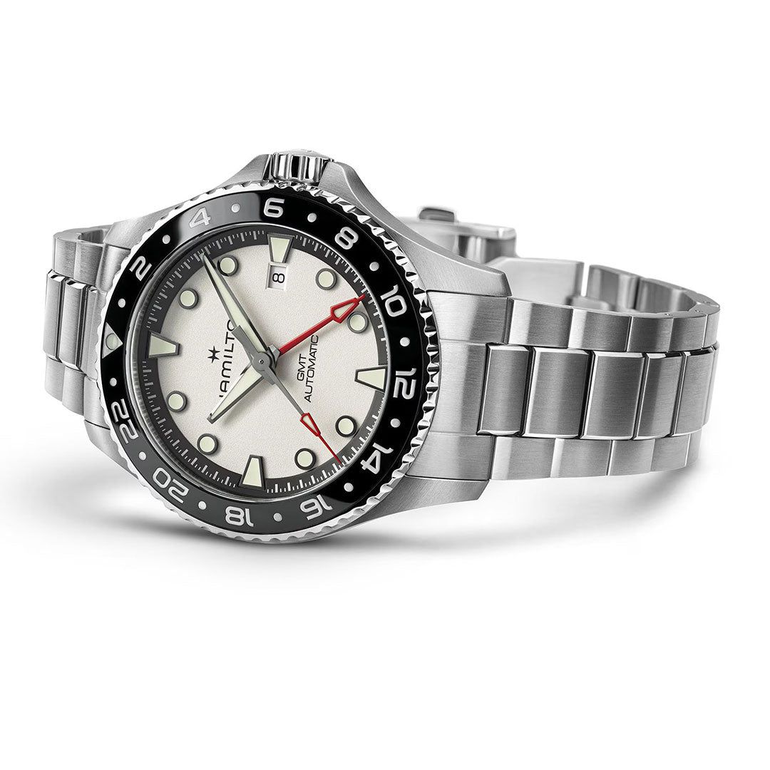 The Hamilton Khaki Navy Scuba Auto GMT 43mm watch by Hamilton Watch features a metal bracelet, black and silver bezel, white dial with red GMT hand, and a date window at 3 o’clock, displayed against a white background.