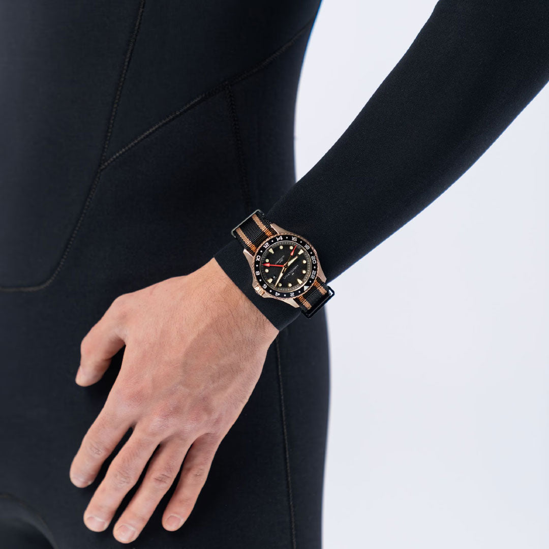 A person in a black wetsuit wears the Hamilton Khaki Navy Scuba Automatic GMT 43mm Bronze Watch with a striped band, highlighting its ability to track multiple time zones. The watch stands out against a plain light background.