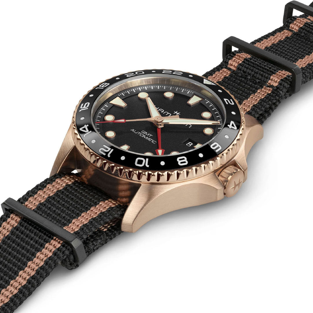 The Hamilton Khaki Navy Scuba Automatic GMT 43mm Bronze Watch by Hamilton Watch features a black and brown striped fabric strap, black bezel with white numerals, black dial, gold markers and hands, plus a red-tipped GMT second hand. Displayed at an angle.