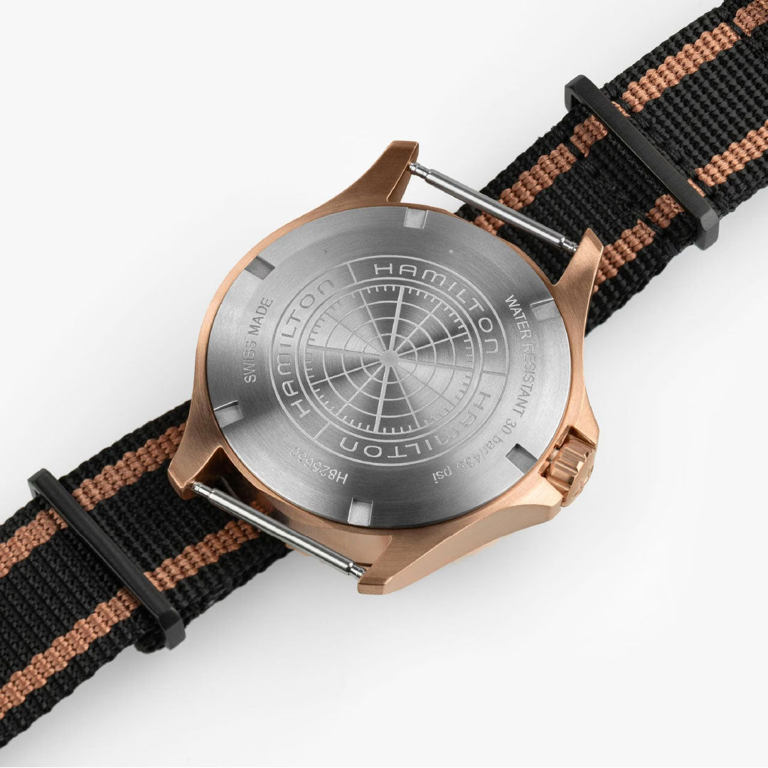 A close-up of the Hamilton Watch Khaki Navy Scuba Automatic GMT 43mm Bronze features a brushed bronze case with engraved details, concentric circular patterns, and a black nylon strap with a tan stripe.