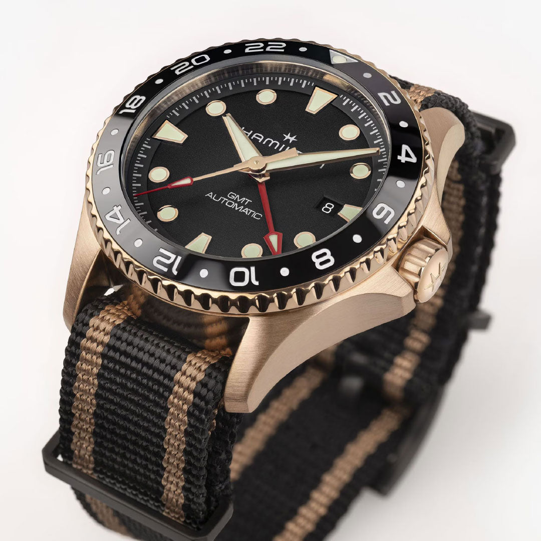 A close-up of the Hamilton Khaki Navy Scuba Automatic GMT 43mm Bronze Watch by Hamilton Watch, featuring a black bezel, black dial with cream markers, and a black/tan striped strap. The date window is at 3 o’clock for easy tracking of multiple time zones.
