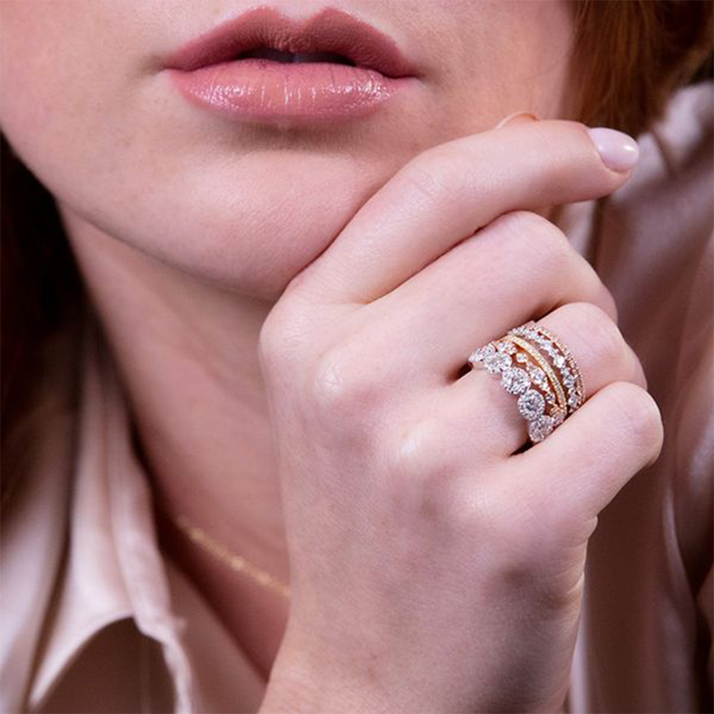 A close-up of a woman's hand touching her chin reveals her adorned fingers, featuring multiple diamond rings and the Hearts On Fire 18K Rose Gold Round Brilliant Diamond Anniversary Ring. Her light skin presents a striking contrast to her pink lips, while she wears a silky, light-colored top that enhances the timeless allure of the scene.