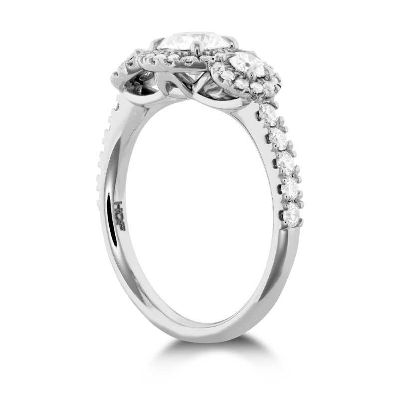 The Hearts On Fire Integrity 18K White Gold Round Brilliant Three-Stone Semi Mount with Diamond Band and Halo Engagement Ring from Hearts on Fire features a central diamond flanked by two smaller diamonds. The elegant band, adorned with a row of smaller diamonds, enhances the ring's sparkle and accentuates its diamond carat weight beautifully.