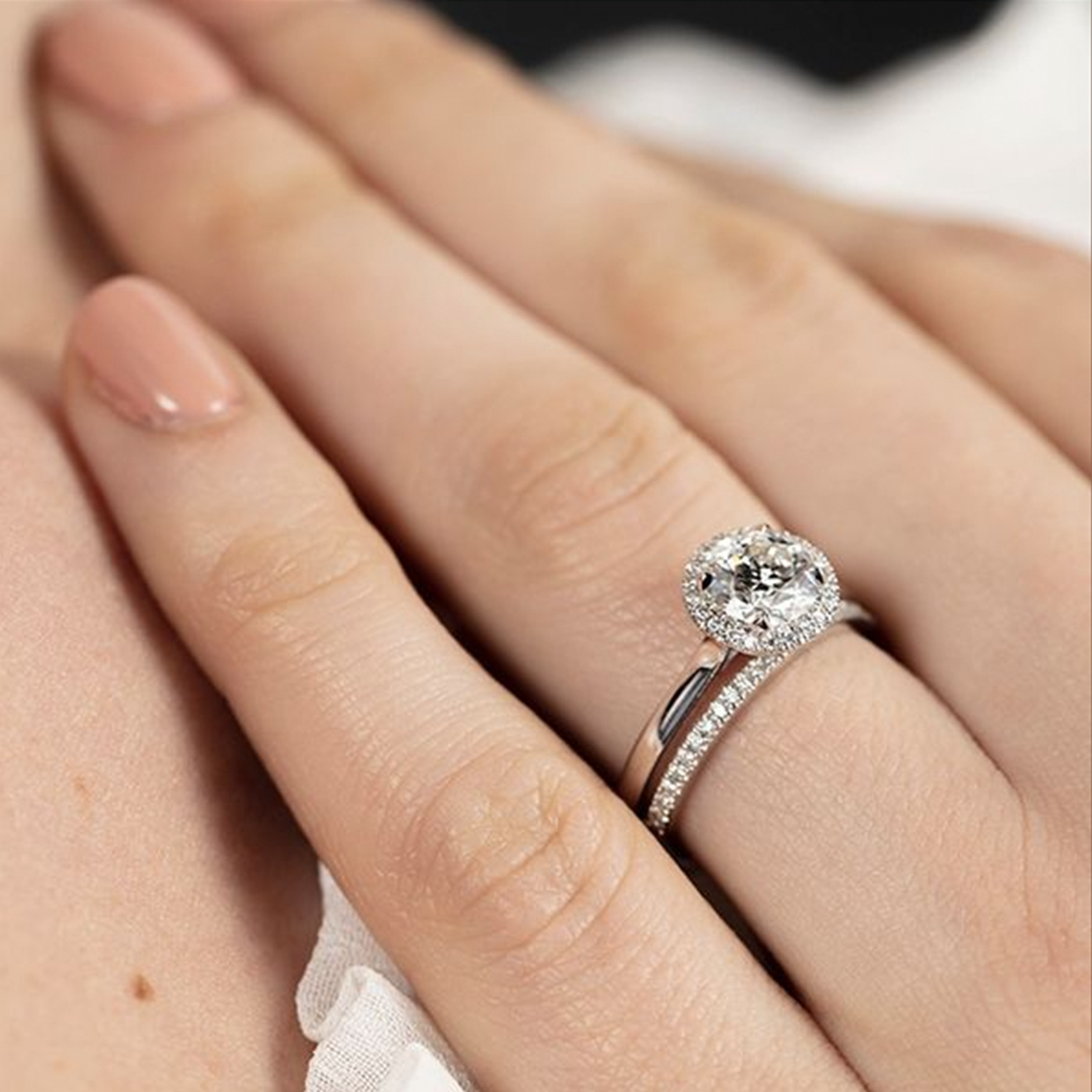 A close-up of a hand adorned with the exquisite Hearts On Fire 18K White Gold Round Brilliant Diamond Halo Engagement Ring. The nails are manicured with light pink polish, highlighting the ring's round diamond center surrounded by smaller diamonds along its elegant white gold band.