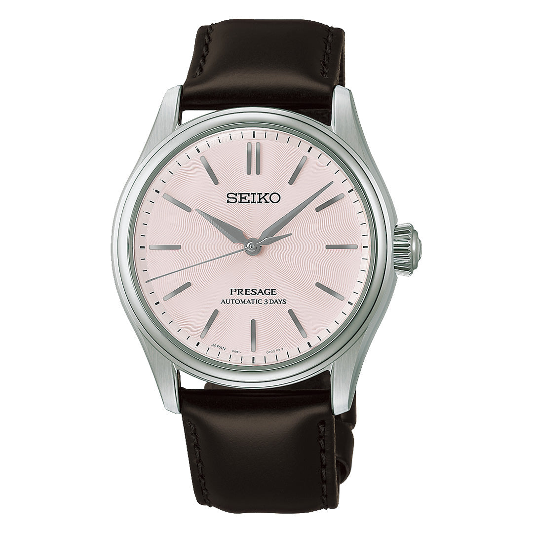 The Seiko Presage Classic Series Automatic 38mm Watch showcases a sophisticated stainless steel case paired with a light pink dial featuring silver hour markers and hands. This elegant timepiece by Seiko is finished with a dark brown leather strap, while the dial is accented with the inscriptions “SEIKO,” “PRESAGE,” and “AUTOMATIC 3 DAYS.”.