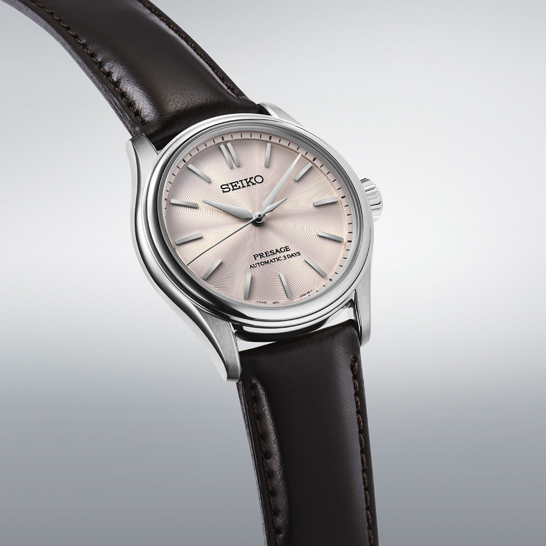 The Seiko Presage Classic Series Automatic 38mm Watch showcases a light pink textured dial with silver indices and a dark brown leather strap, all elegantly presented against a gray gradient background.