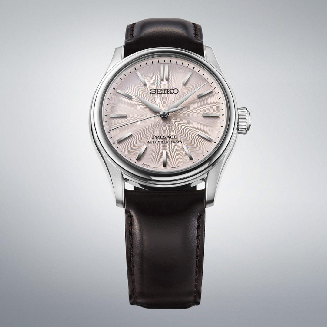 The Seiko Presage Classic Series Automatic 38mm Watch by Seiko showcases a light pink dial with silver hour markers and a black leather strap, beautifully presented against a neutral gray background.