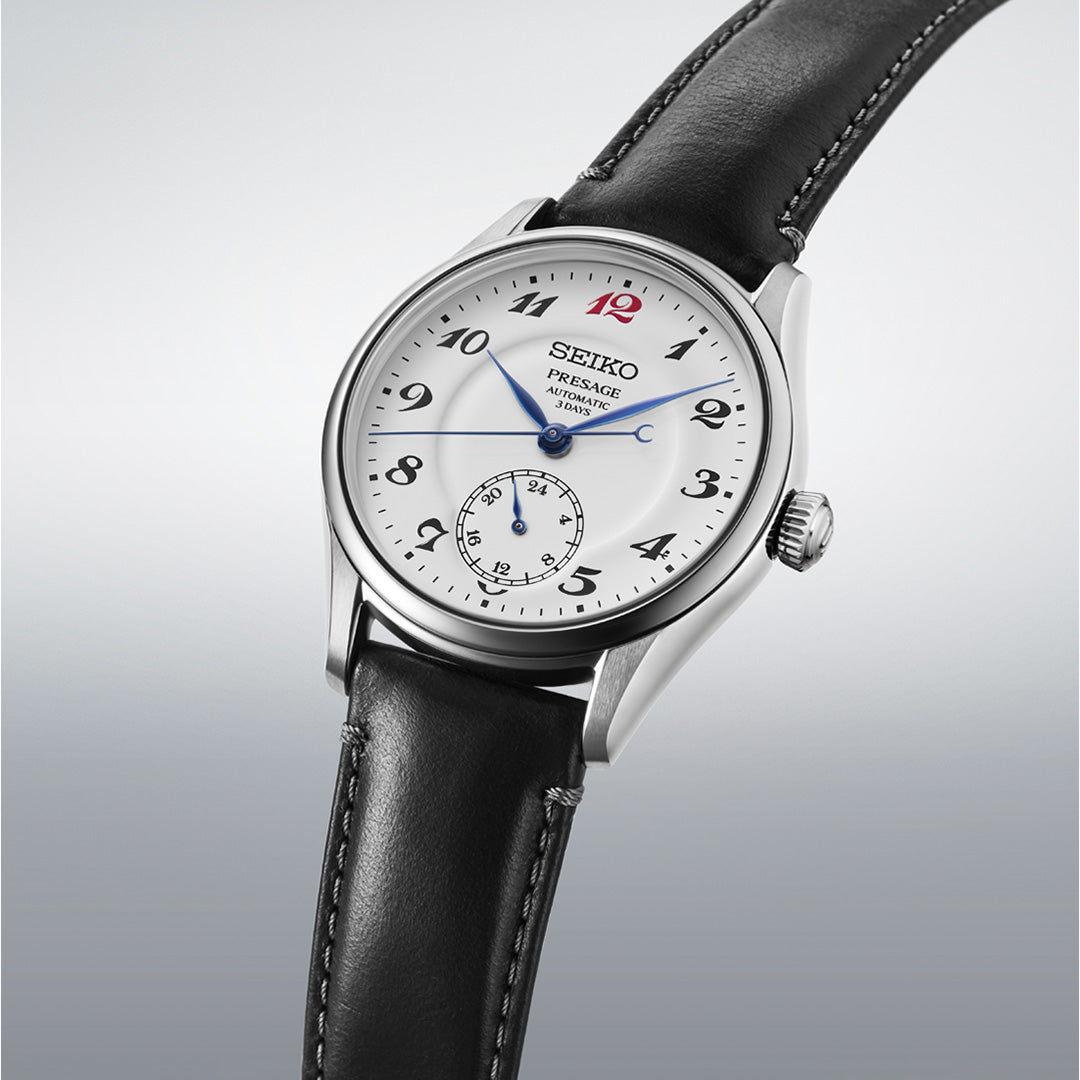 The Seiko Presage Classic Series Automatic 39.6mm Watch by Seiko features a white dial with black Arabic numerals, blue hands, a small seconds subdial at 6 o’clock, a bold red “12” at the top, and comes with a black leather strap. It is shown against a light gray background.