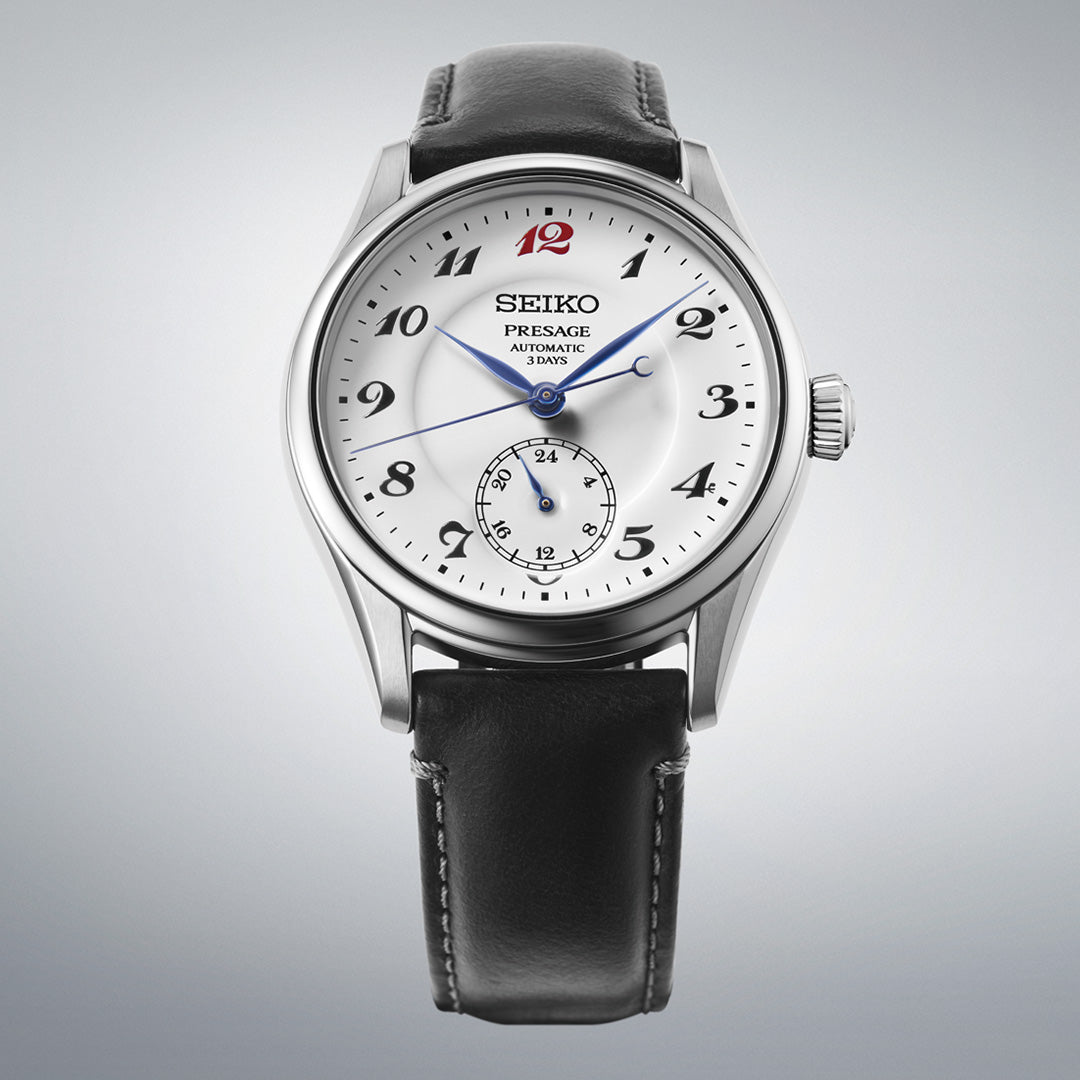 The Seiko Presage Classic Series Automatic 39.6mm Watch by Seiko features a refined white dial adorned with black Arabic numerals, bold blue hands, and a distinctive red 12 o’clock marker. It includes a subdial at the 6 o’clock position, a power reserve indicator at 9 o’clock, and comes complete with a classic black leather strap.