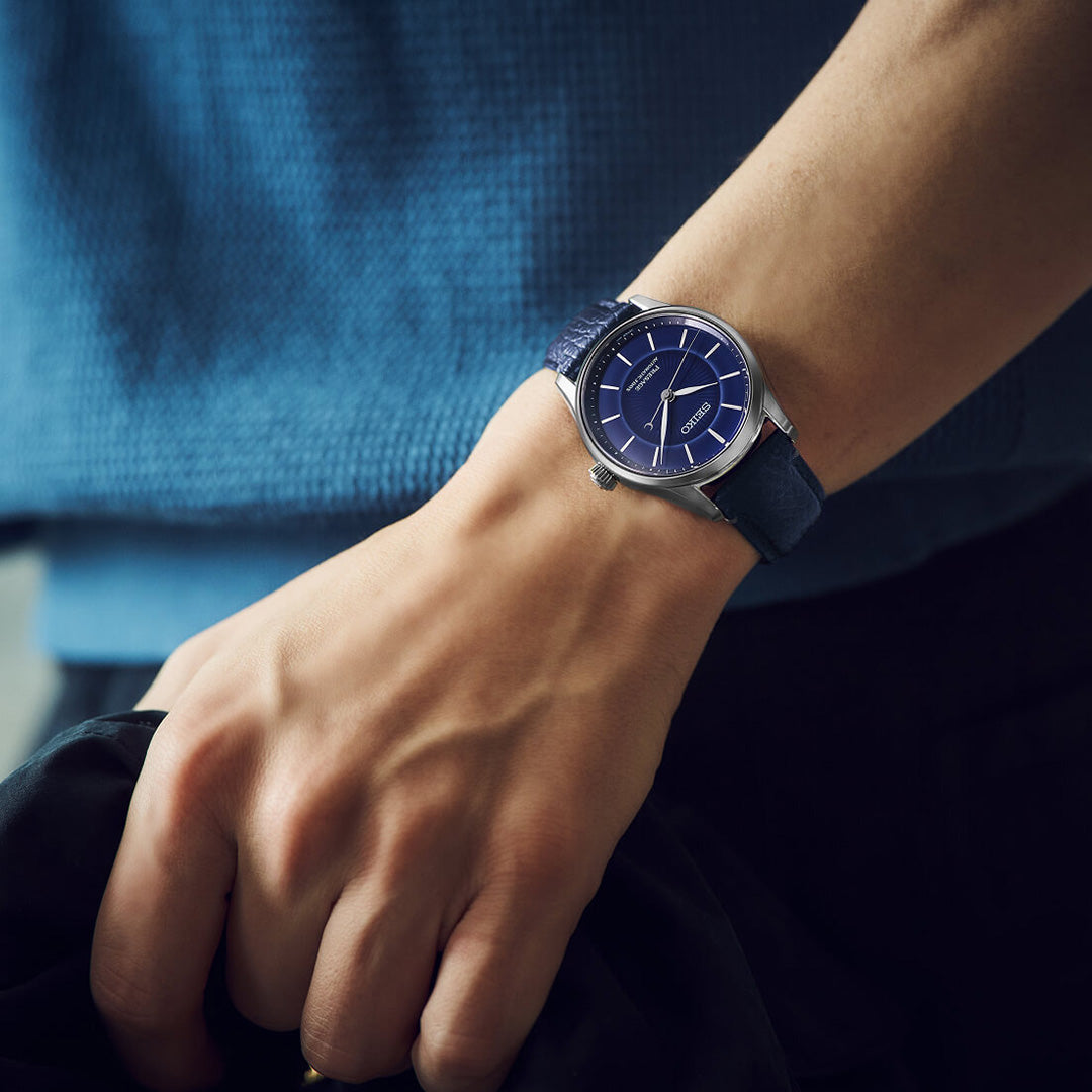 A person wearing the Seiko Presage Classic Series 145th Anniversary Automatic 39.6mm Watch— a limited edition of 1,500 pieces by Seiko— with a stainless steel round case and leather strap, stands with their hand relaxed, dressed in a blue textured top.