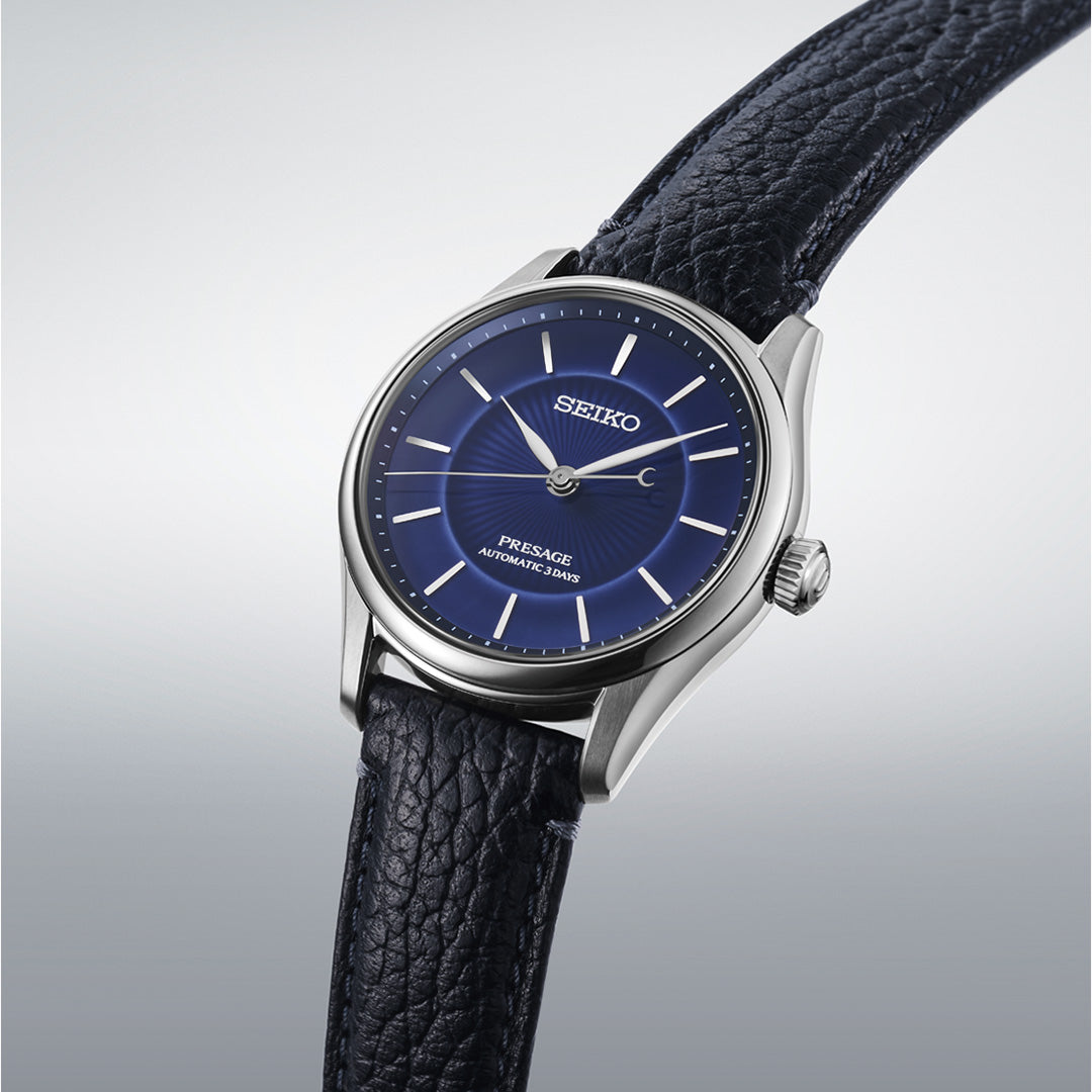 A Seiko Presage Classic Series 145th Anniversary Automatic 39.6mm Watch, limited to 1,500 pieces, features a striking blue dial with silver hour markers and a black textured leather strap. Displayed at an angle against a gradient gray background, this timepiece showcases Seiko’s renowned blend of timeless elegance and modern craftsmanship.